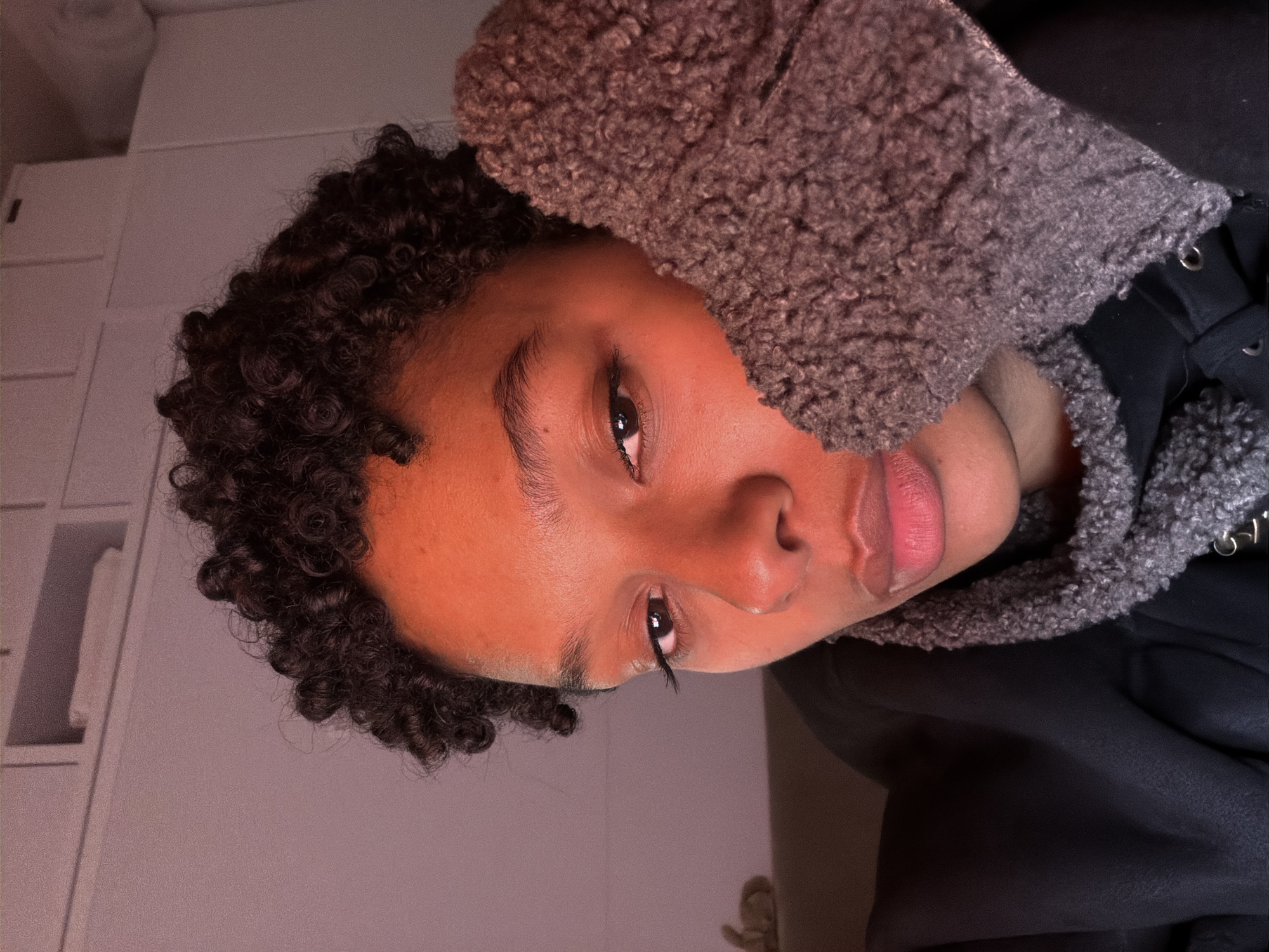 A close-up, slightly rotated image shows a person with dark curly hair, partially obscured by a gray, textured hood. The person's skin appears orange in the lighting. A portion of a gray or white cabinet is visible in the upper left background. The image's location is undetermined.