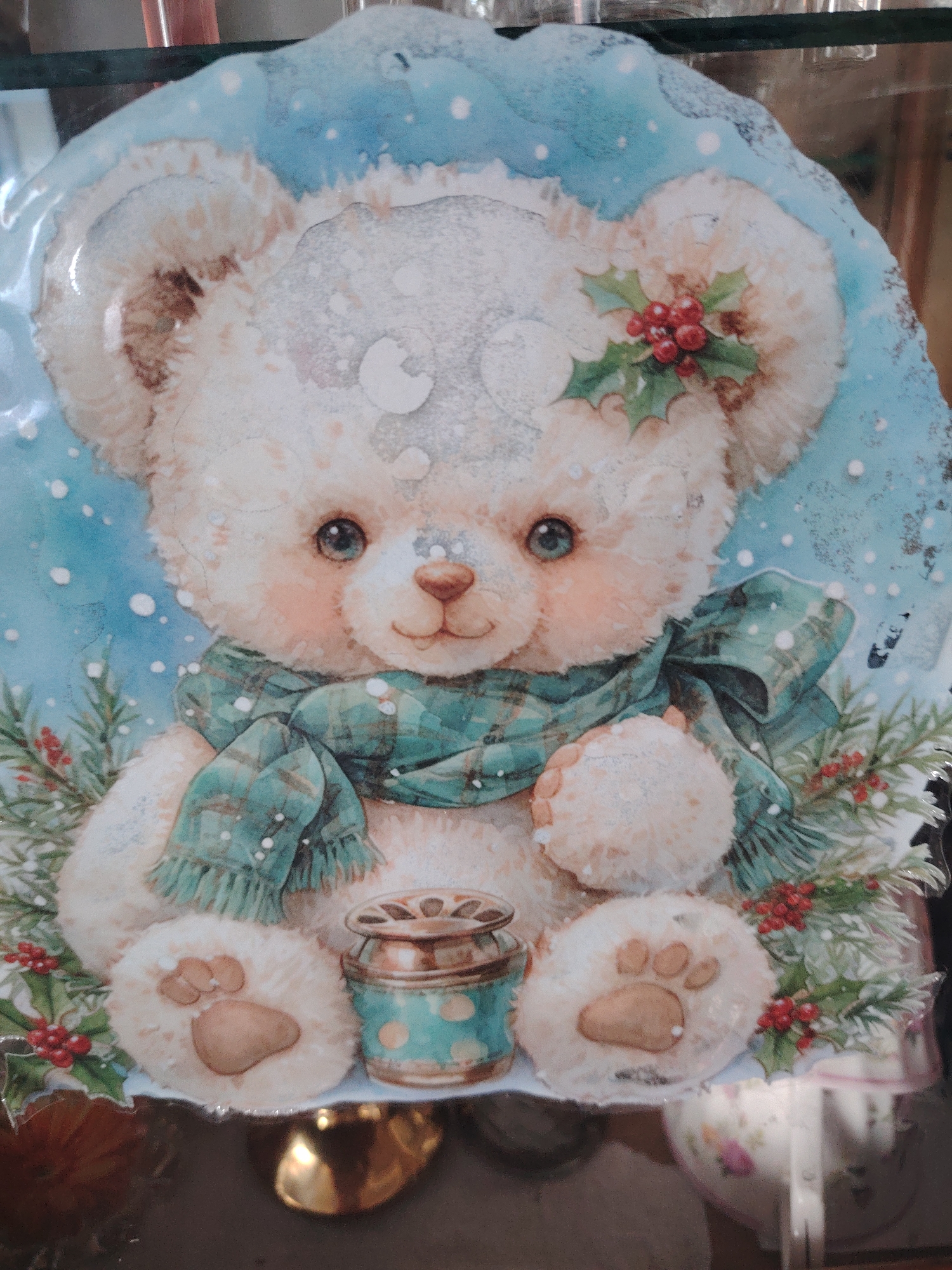 The image displays a charming illustration of a light-colored teddy bear, rendered in a soft, watercolor-like style, set against a snowy blue background. The bear has large, expressive blue eyes, a small brown nose, and rosy cheeks. Its fluffy fur is depicted with subtle shading and is dusted with white snowflakes.
The bear is adorned with a plaid scarf in shades of teal and green, tied around its neck. A sprig of holly with bright red berries and green leaves is tucked behind its left ear, and similar holly sprigs, along with evergreen branches, frame the bear from below.
Next to the bear's right paw, there's a small, decorative cylindrical jar or container with a metallic gold-colored top and a teal body featuring gold polka dots. The bear's paws show distinct, brown paw pads.
The overall scene evokes a festive, winter, or Christmas theme. The item itself appears to be a decorative plaque or plate, given its slightly irregular contoured shape and glossy surface, which reflects some light and elements from its surroundings (e.g., a golden object at the bottom). There is no discernible activity taking place, as it is a static artwork. No text is visible in the image.