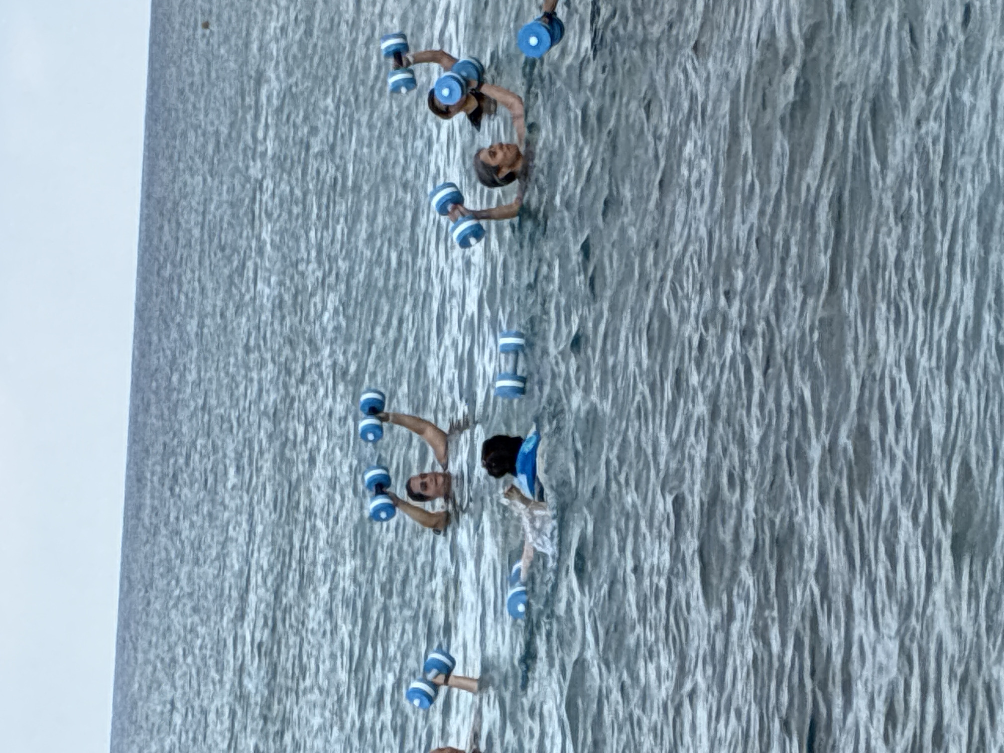 In Alanya, Turkey, a group of individuals is engaged in water aerobics in the ocean. The water is a moderate blue-gray, with small waves. Several people are visible, mostly from the chest up, holding blue and white dumbbells. One person in the foreground on the left appears to be wearing a life jacket. The sky is overcast and a light gray. The scene suggests an outdoor fitness class taking place in the sea.