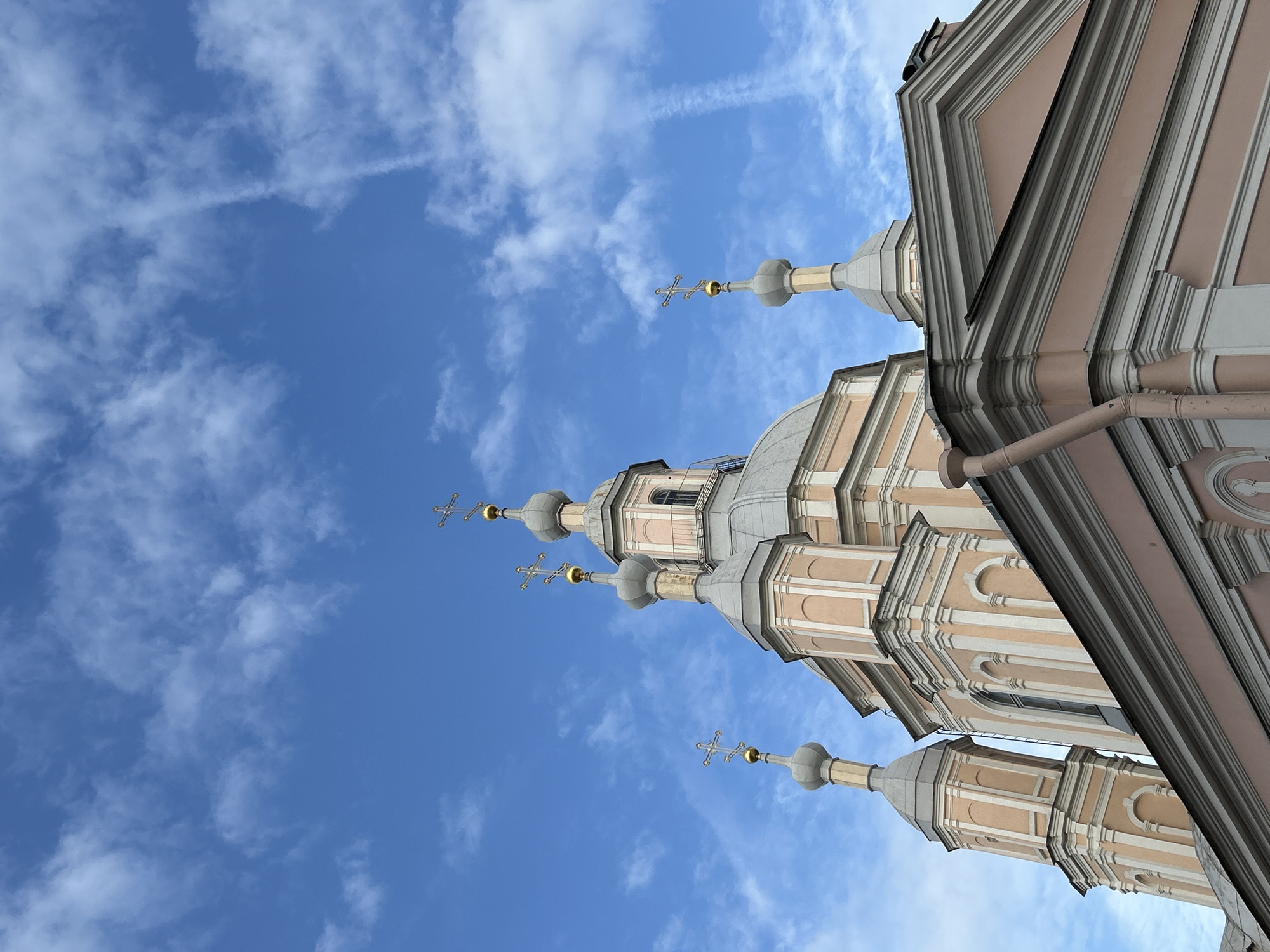 The image shows a low-angle view of the Smolny Convent in Saint Petersburg, Russia. The architecture includes multiple domes topped with crosses, set against a blue sky with light clouds. The building's color scheme features light pink and gray. A portion of the building's corner is visible, showing architectural details like eaves and decorative moldings. No people are visible in the frame.