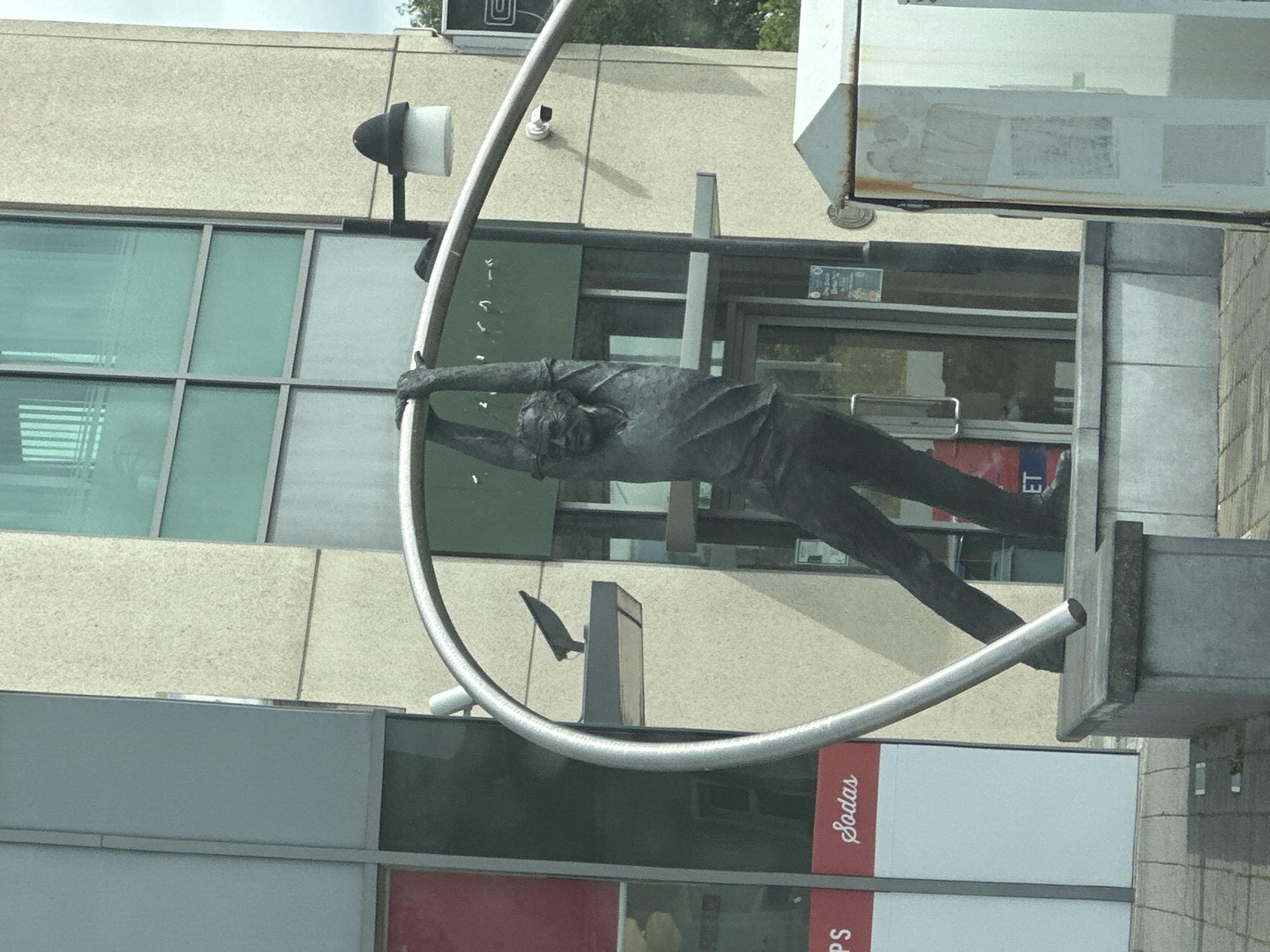A dark bronze statue of a man hanging from a metal archway is displayed in front of a modern building with large windows. The man is depicted with his arms extended and gripping the arch, with his body in a bent position. The building appears to be a commercial establishment, with signage visible on the windows, including "PS" and "Sodas." The statue is situated on a concrete platform, which is placed on a paved area. In the background, a lamppost with a dark cap is visible, and a security camera is mounted on the building. The overall scene suggests an urban public art installation. This statue is known as the "George Bernard Shaw Sculpture" and is located in Carlow town, County Carlow, Ireland.