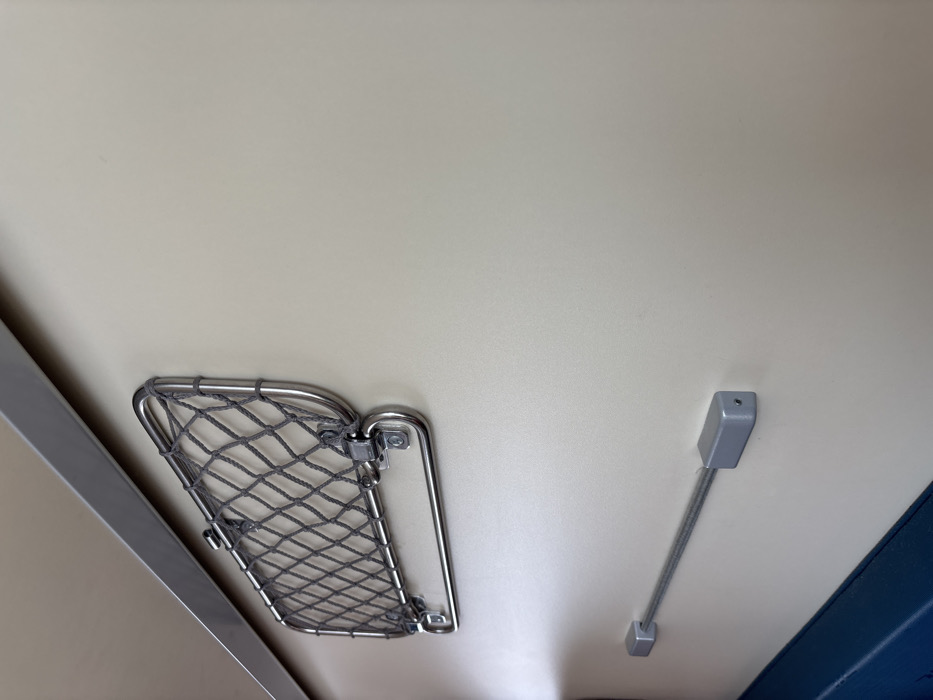 The image depicts an interior view of a compartment, likely within a modern transport vehicle such as a train. The primary surface is a smooth, light beige or cream-colored wall that occupies most of the frame. Along the upper left edge of the wall, a polished metallic trim piece, appearing to be stainless steel or aluminum, runs horizontally.
Mounted on the wall in the upper-left quadrant is a polished stainless steel frame containing a gray, coarse-mesh net, serving as a luggage or storage net. Directly below this net, a separate, hinged stainless steel bar is attached horizontally.
Below the luggage net and slightly to its right, another horizontal utility fixture is present. This consists of two rectangular, silver-colored end mounts attached to the wall, between which a thin, coiled or flexible silver-colored bar extends.
At the bottom-left edge of the frame, a small section of a dark blue surface is visible, consistent with the top edge of an upholstered seat or bench. No individuals, actions, or specific interactions are present in the scene. The lighting is even, originating from the left. The image provides no visual information to determine a specific geographical location within Ivanovka, Russia or any other city.