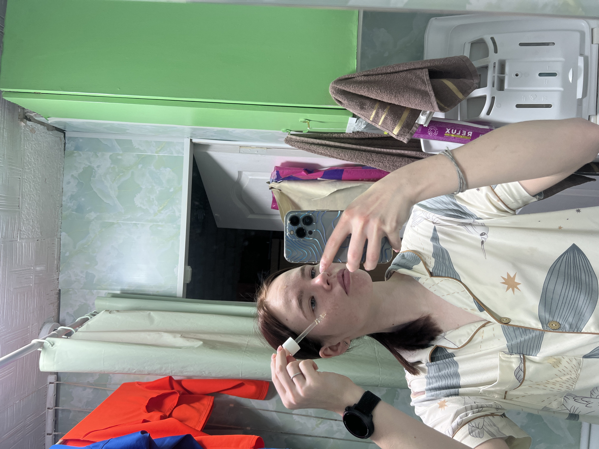 A female is taking a selfie in a bathroom. She is holding a dropper with a liquid product near her face. She is wearing pajamas and a smartwatch. The bathroom has green cabinets, white tiled walls, and a shower curtain rod. Clothing is draped over the curtain rod. A brown towel and cleaning supplies are stored above the cabinets. The location is in Chaikovsky, Russia.