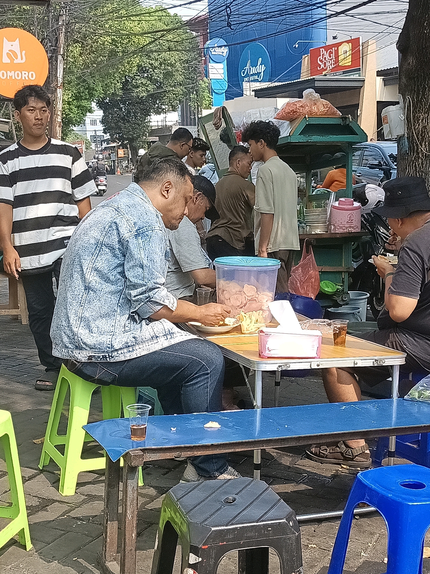 This image captures a street scene in South Jakarta, Indonesia, likely near a bustling commercial area. In the foreground, several individuals are gathered around small tables, some consuming food or drinks. A man in a denim jacket sits on a bright green plastic stool, his attention focused on food on a small table. Adjacent to him, another man in a dark t-shirt and bucket hat is also eating. The tables are laden with various items, including plastic containers filled with snacks, a tissue box, and glasses of beverages.
Several other people are visible in the background. A young man in a black and white striped shirt stands near a circular orange sign with a cat logo. Other men are seen standing and interacting, suggesting a social gathering or a break from work.
The environment appears to be outdoors, possibly along a sidewalk or a small market area. The background reveals a street with passing vehicles, buildings with signage, and abundant overhead electrical wires, characteristic of urban Indonesian settings. Trees provide some shade, and the ground is paved with interlocking bricks. The overall atmosphere suggests a casual, everyday scene in South Jakarta.