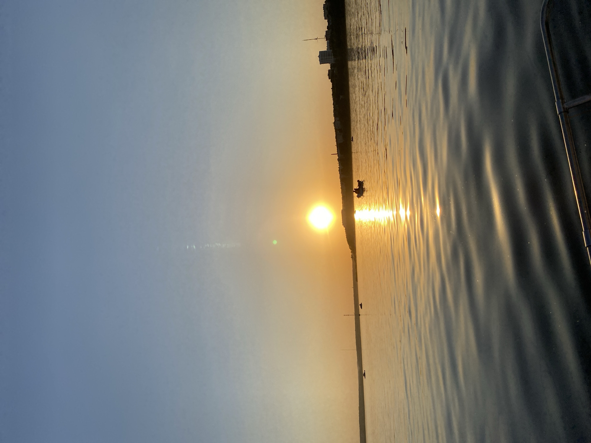 The image shows a sunset over a body of water, likely a river or lake, in Izhevsk, Russia. The sun is positioned on the horizon, casting a bright reflection on the water's surface. The sky transitions from a pale blue to an orange hue near the horizon. A few buildings are visible in the distance along the shoreline. Two small boats with people are present on the water, silhouetted against the sunlight. The foreground of the image shows a part of a boat.