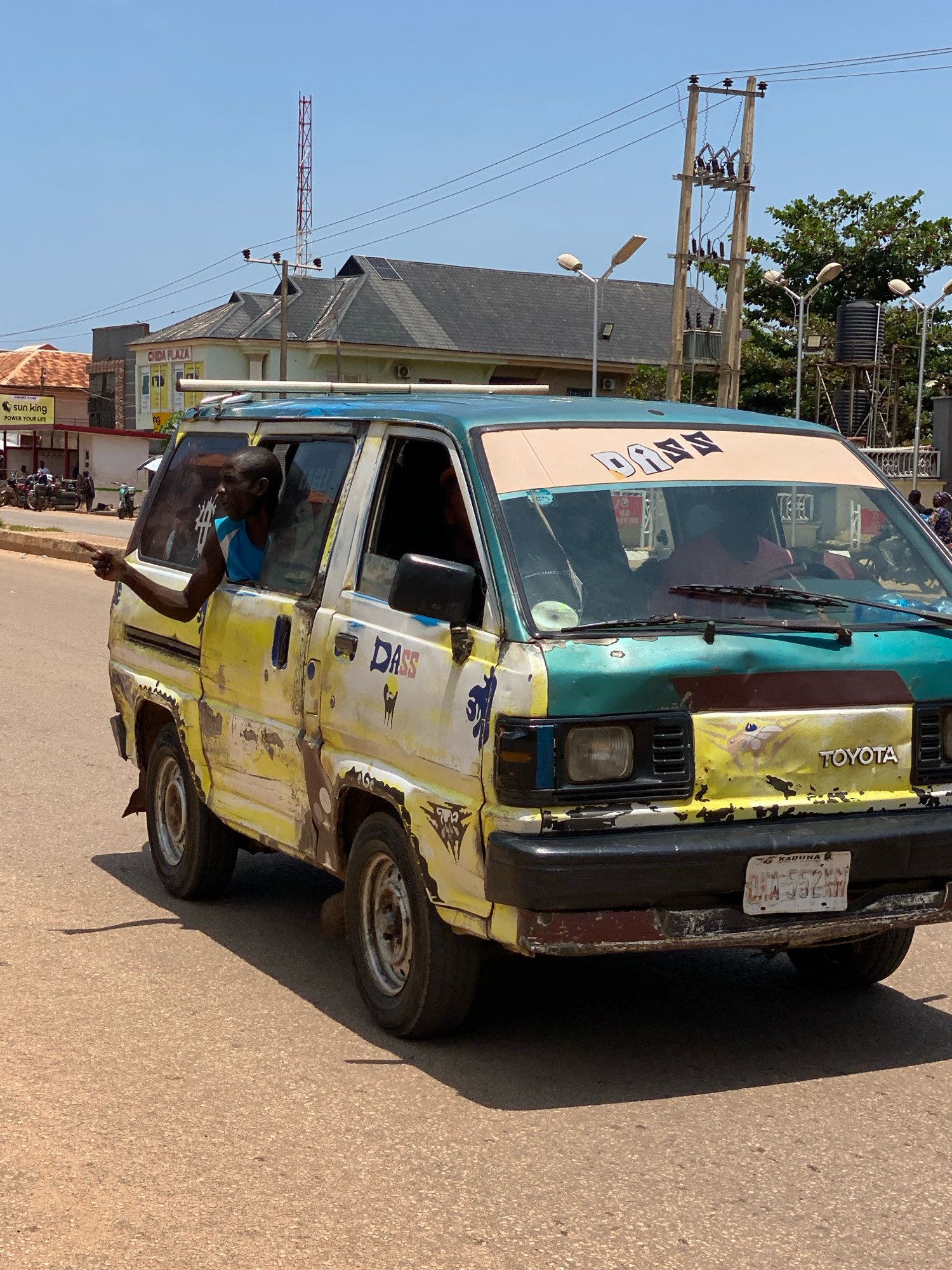 A worn, yellow and teal Toyota Hiace minivan, with the word "DASS" painted in large letters across its windshield, travels down a dusty, paved road in Sabon Tasha, Nigeria. A Black man leans out the passenger-side window, pointing his left index finger forward, seemingly indicating a direction. His right arm is also visible resting on the window frame. The van's exterior is heavily weathered, with peeling paint, rust, and various decals and markings, including "TOYOTA" emblazoned on the front. The license plate, partially obscured, reads "OKA 552 AM KADUNA." In the background, buildings with peaked roofs, solar panels, and utility poles with power lines are visible under a clear, bright sky. The overall environment suggests a busy urban or suburban street scene in Nigeria.