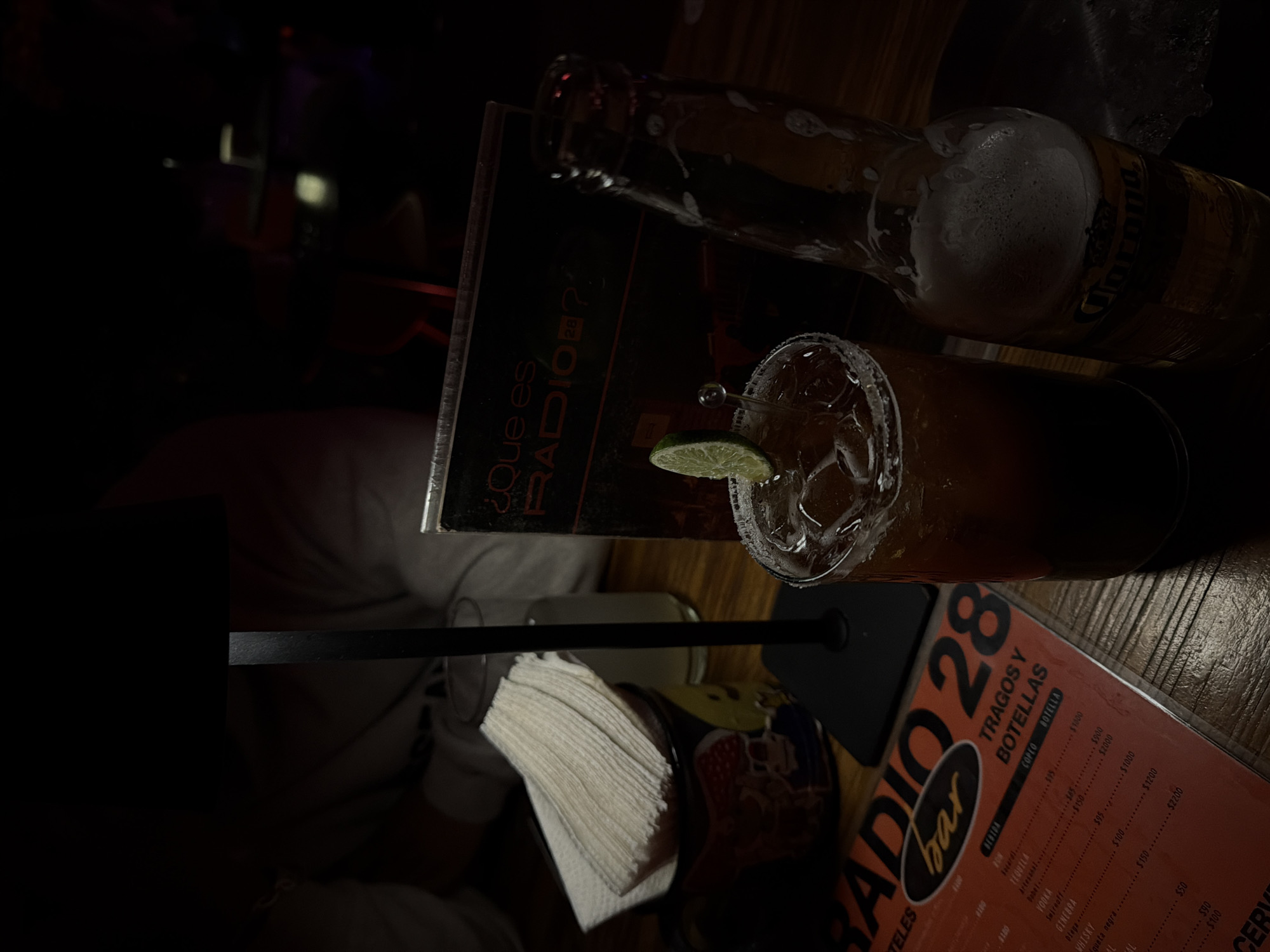 The image captures a table at Radio Bar 28 in San Andrés Cholula, Mexico. A partially consumed bottle of Corona beer rests near a glass of margarita, garnished with a lime wedge. A folded menu from Radio Bar 28 displays the prices of drinks and other beverages. A stack of napkins sits nearby. The overall lighting is dim. A person's arm is visible to the left of the napkins.