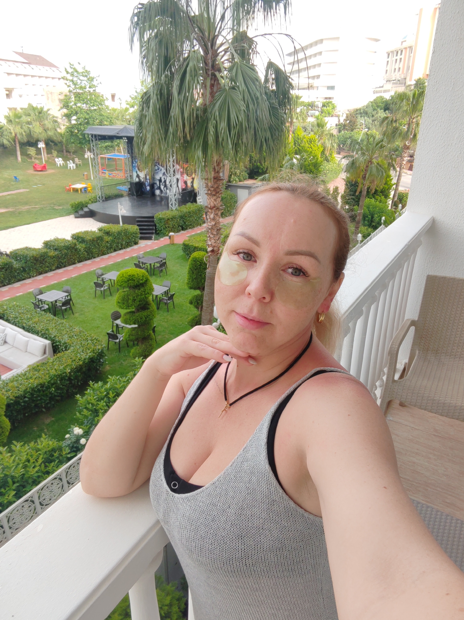 A selfie shot depicts a blonde-haired woman on a balcony overlooking an outdoor recreation area. She has green under-eye patches on and wears a gray tank top with a black necklace. The woman is leaning against the white balcony railing, her hand resting on her chin. Behind her, a well-manicured lawn is visible, featuring topiaries, tables with chairs, and a white outdoor sofa. In the distance, there's a stage with a black roof and painted backdrop. Beyond the lawn, there are palm trees and multi-story buildings. The location is identifiable as Alanya, Turkey, due to the architectural style, landscaping, and the presence of palm trees characteristic of the Mediterranean region.