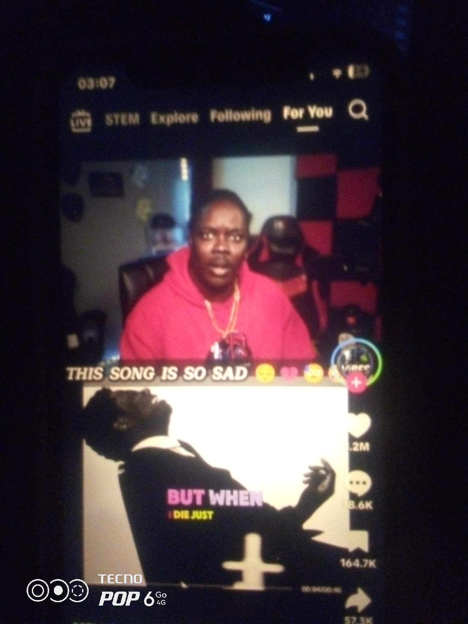 The image depicts a smartphone screen displaying a video content platform, likely TikTok or a similar social media app. The time displayed at the top left is "03:07". A person wearing a red hoodie and a gold chain is visible in the upper portion of the screen, reacting to a video. Below this person, text overlays read "THIS SONG IS SO SAD" accompanied by emojis. The lower portion of the screen shows a silhouette of a person with the text "BUT WHEN I DIE JUST" superimposed. On the right side of the screen, interaction icons for likes, comments, and shares are visible, with numerical counts associated with them. The bottom left corner of the image shows the branding "TECNO POP 6 Go 4G".
The prompt specifies to include the location in the city of Howayi, Nigeria. However, there is no visual information within the image that indicates the specific location of Howayi, Nigeria. The image solely captures a screen display on a mobile device.