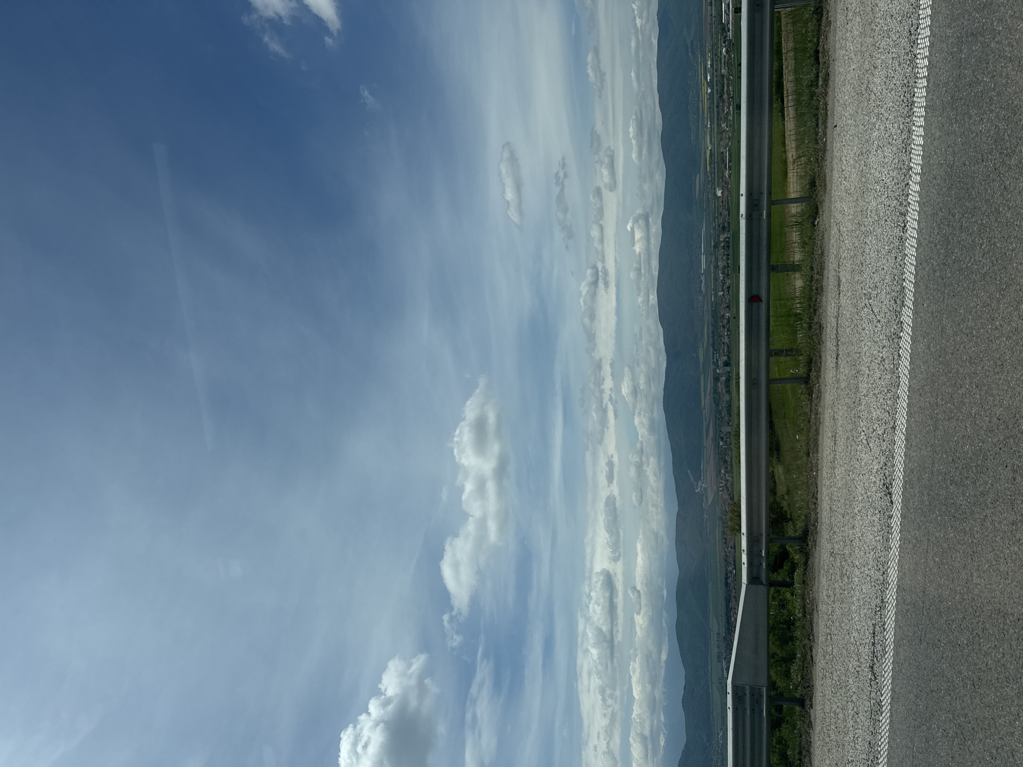 The image depicts a view from a roadway, possibly a highway or freeway. A guardrail is present between the road surface and a grassy embankment. In the distance, mountains are visible under a sky with scattered clouds. The location is unknown.