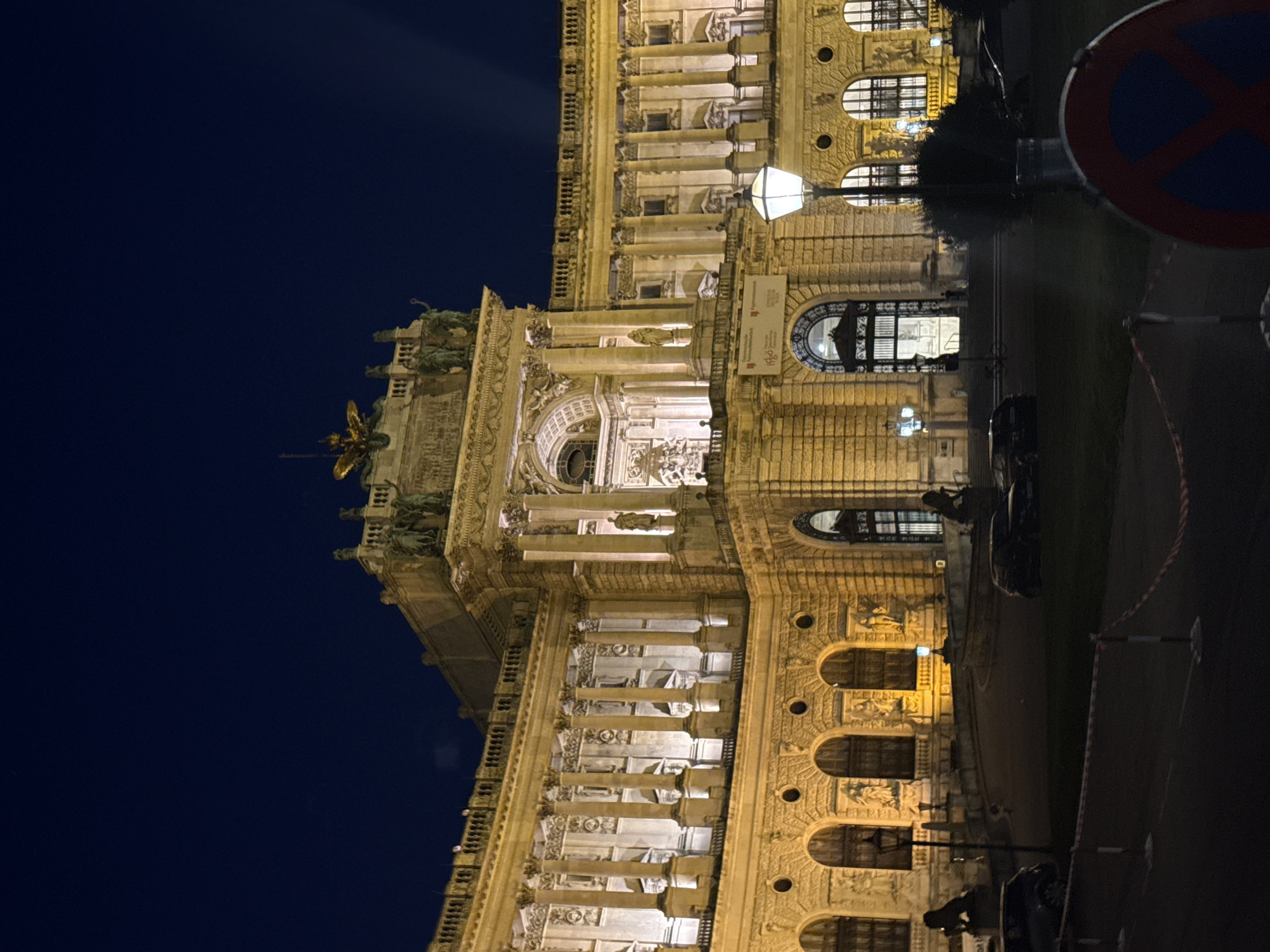 The image depicts a grand, illuminated building at night in Vienna, Austria, identified as the Hofburg Palace. The architectural style is predominantly Baroque, characterized by ornate facades, numerous columns, arched windows, and sculptural decorations. The building is warmly lit by artificial lights, highlighting its intricate details and casting a golden hue against the dark night sky. Several statues and architectural embellishments are visible on the upper levels and facade. In the foreground, a lamppost with a lit lantern stands prominently. Two cars are parked on the grass in front of the building, and the area is cordoned off with red and white striped tape. A "no parking" sign is partially visible in the bottom right corner. The overall scene conveys a sense of historical grandeur and nocturnal ambiance.