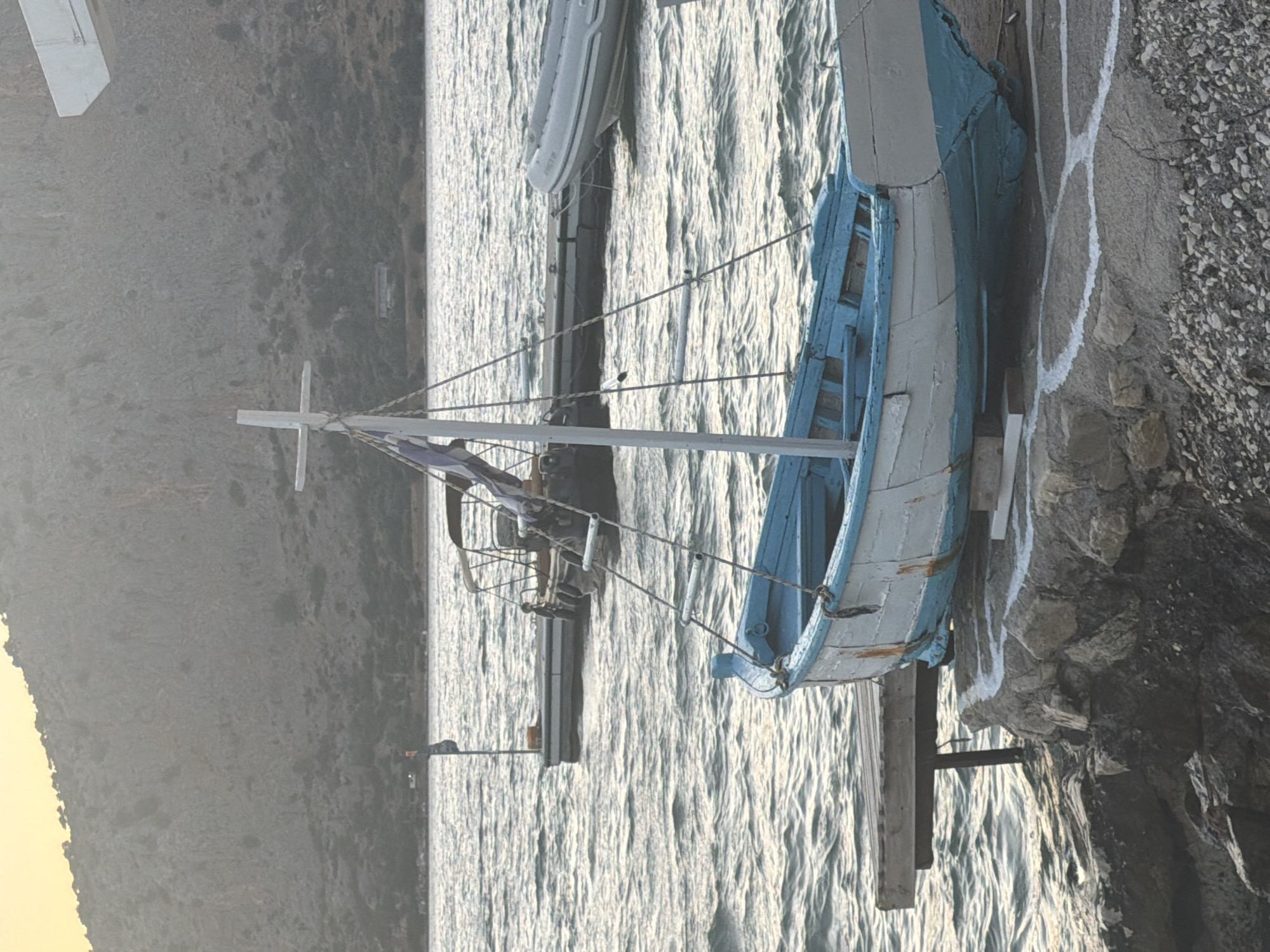 The image depicts a serene coastal scene in the Municipal Unit of Alyzia, Greece. In the foreground, a weathered wooden boat with a white hull and a blue trim rests on a rocky shore. A simple wooden jetty extends into the rippling water to the left of the boat. In the middle ground, several other vessels are moored or docked, including an inflatable boat and a larger boat with a canopy. The water is calm and reflects the hazy, pale yellow light of the sky, suggesting it is either dawn or dusk. In the background, a steep, rocky mountain covered in sparse vegetation rises from the coastline. The overall impression is one of quiet tranquility in a Mediterranean setting.