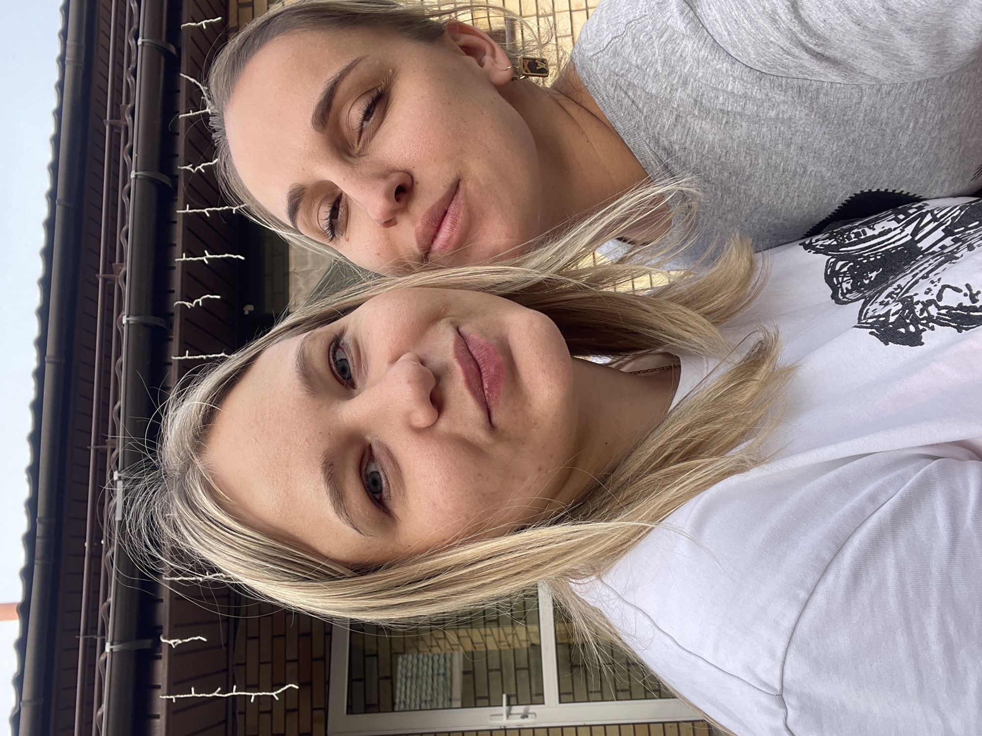 Two blonde women are posing for a selfie outdoors. The woman on the bottom is wearing a white t-shirt with a black and white design on the sleeve, and the woman on top is wearing a gray t-shirt. The background includes a brick building with a window and decorative lights strung along a metal railing. Based on metadata, this photo was taken in Chaikovsky, Russia.