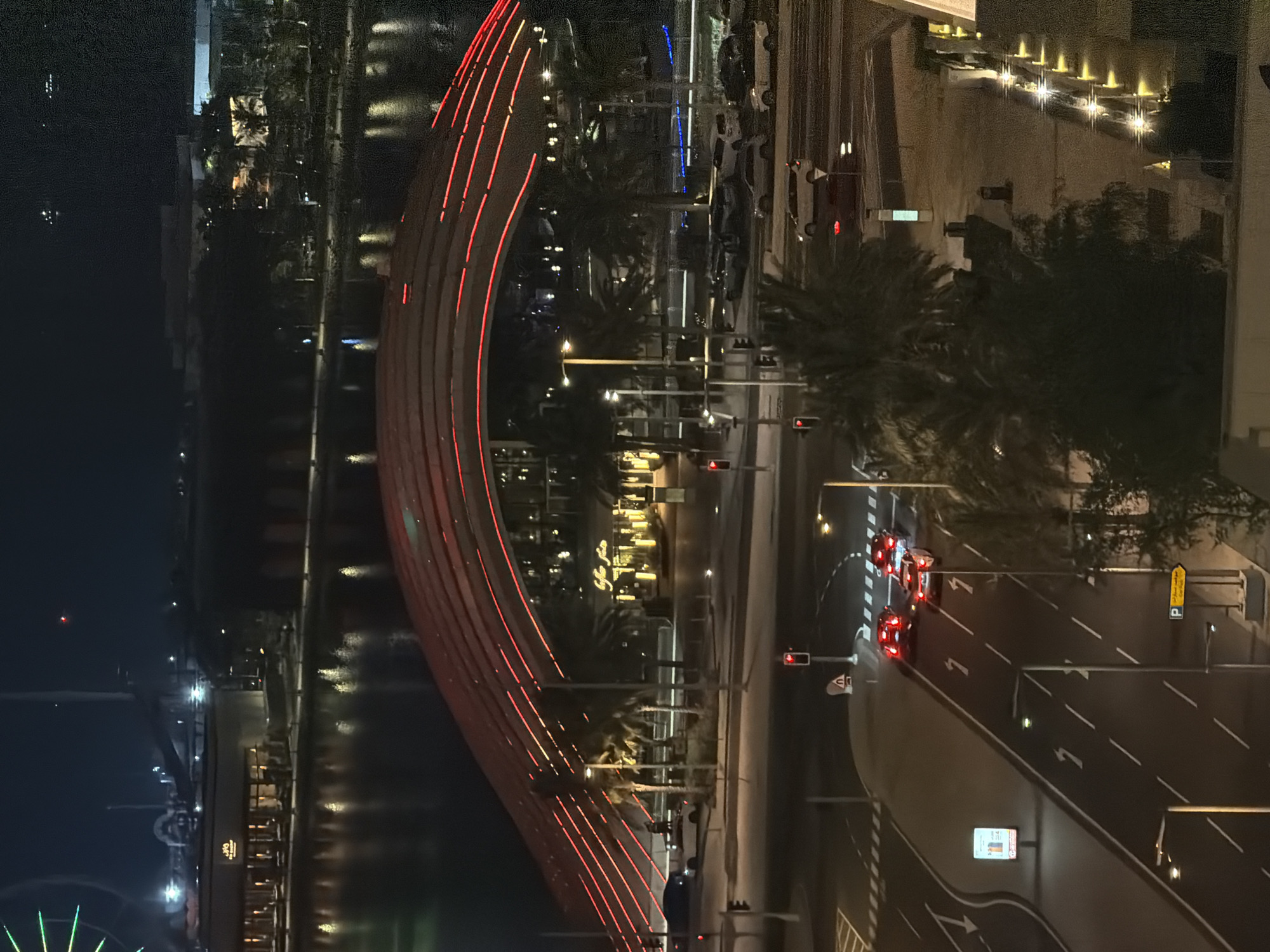 A high-angle, full outdoor shot captures a cityscape at night. The scene is dominated by a large, arched bridge illuminated with red lights, spanning over a body of water. Several buildings are visible on the far side of the water, with windows lit up. In the foreground, a multi-lane road runs horizontally across the image, with several cars present. Traffic lights are visible, with some showing red. Streetlights and palm trees line the road. The overall ambiance is one of urban nighttime activity.