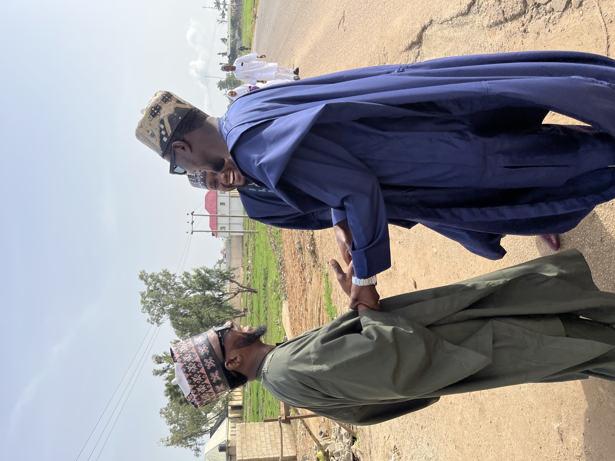Two men in traditional Hausa attire are engaged in a handshake on a cracked asphalt road. The man on the left is wearing a dark green robe and a patterned hat, while the man on the right is wearing a dark blue robe and a similar patterned hat. Both men are wearing sunglasses and have facial hair. In the background, there is a building with a red roof, green vegetation, and other people in white clothing further down the road. The location appears to be on the outskirts of the city of Jos, Nigeria, based on the style of dress and architecture.