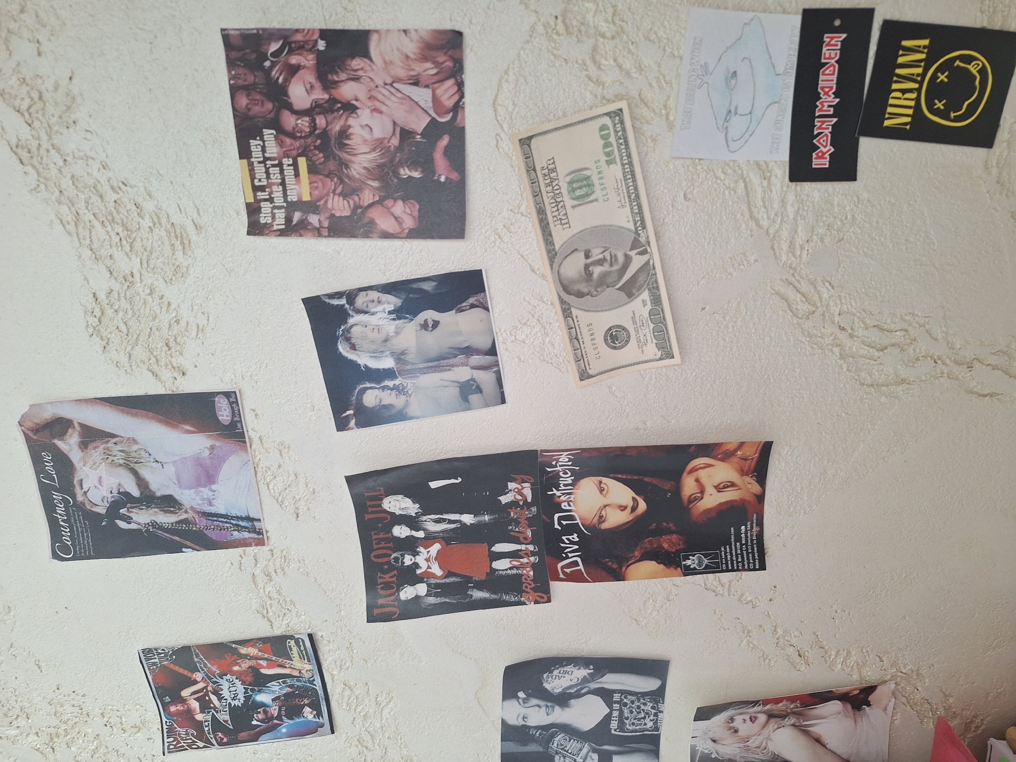 The image shows a collection of stickers and a hundred-dollar bill attached to a textured, off-white wall. The stickers feature various band names and album covers: "Stop it, Courtney. That joke isn't funny anymore", "Courtney Love" album cover, "Jack Off Jill", "Diva Destruction", "Queens of the Stone Age", "Iron Maiden", and "Nirvana" logo, along with a drawing of a cartoon dog. The items are haphazardly arranged on the wall. There is no indication of Panagyurishte, Bulgaria, or any specific location within a city.