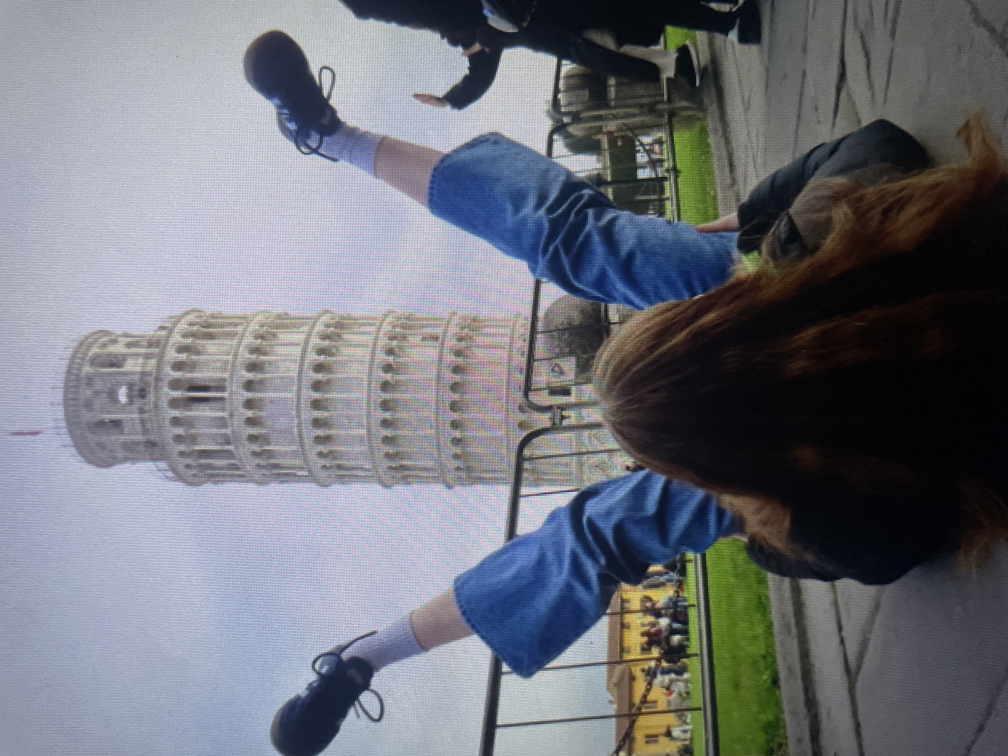A person is lying on their back on a paved surface with their legs raised in the air, appearing to pose for a photo with the Leaning Tower of Pisa in the background. The person is wearing blue jeans, white socks, and dark shoes. The Leaning Tower of Pisa is a tall, cylindrical building with a distinctive tilt, constructed of white stone. A metal fence runs across the foreground. In the background, there are other people and buildings. The sky is overcast. The scene is set in the city of Kołobrzeg, Poland, at the Leaning Tower of Pisa.