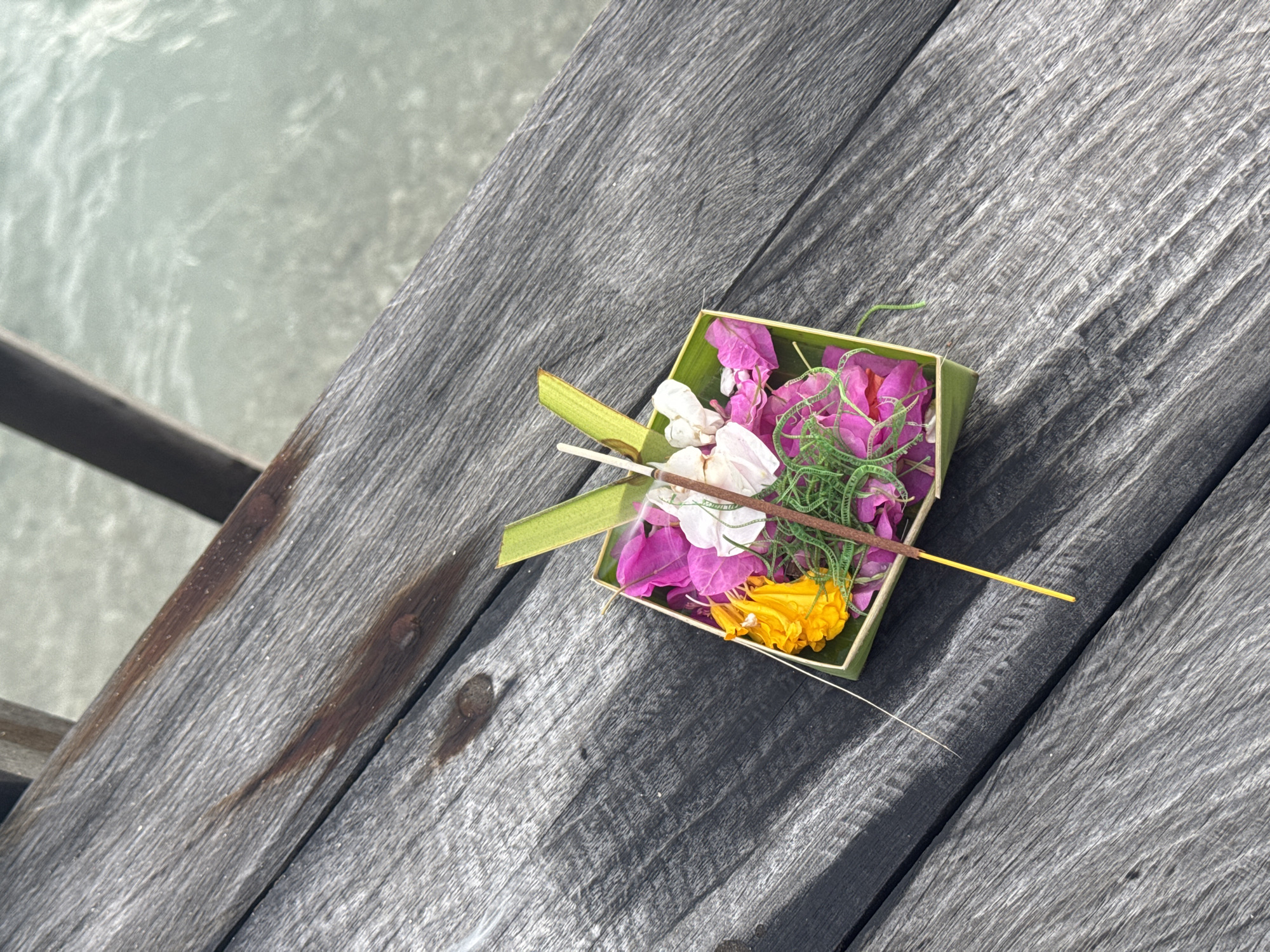 An eye-level, medium shot shows an offering on a gray wood pier at Nusa Penida, Indonesia. The offering, contained in a square green container, includes pink, white, and yellow flowers along with green sprigs. Two pale sticks and a yellow incense stick also reside in the container. Gray wood planks make up the pier. Beyond the pier, out of focus, is the sea.