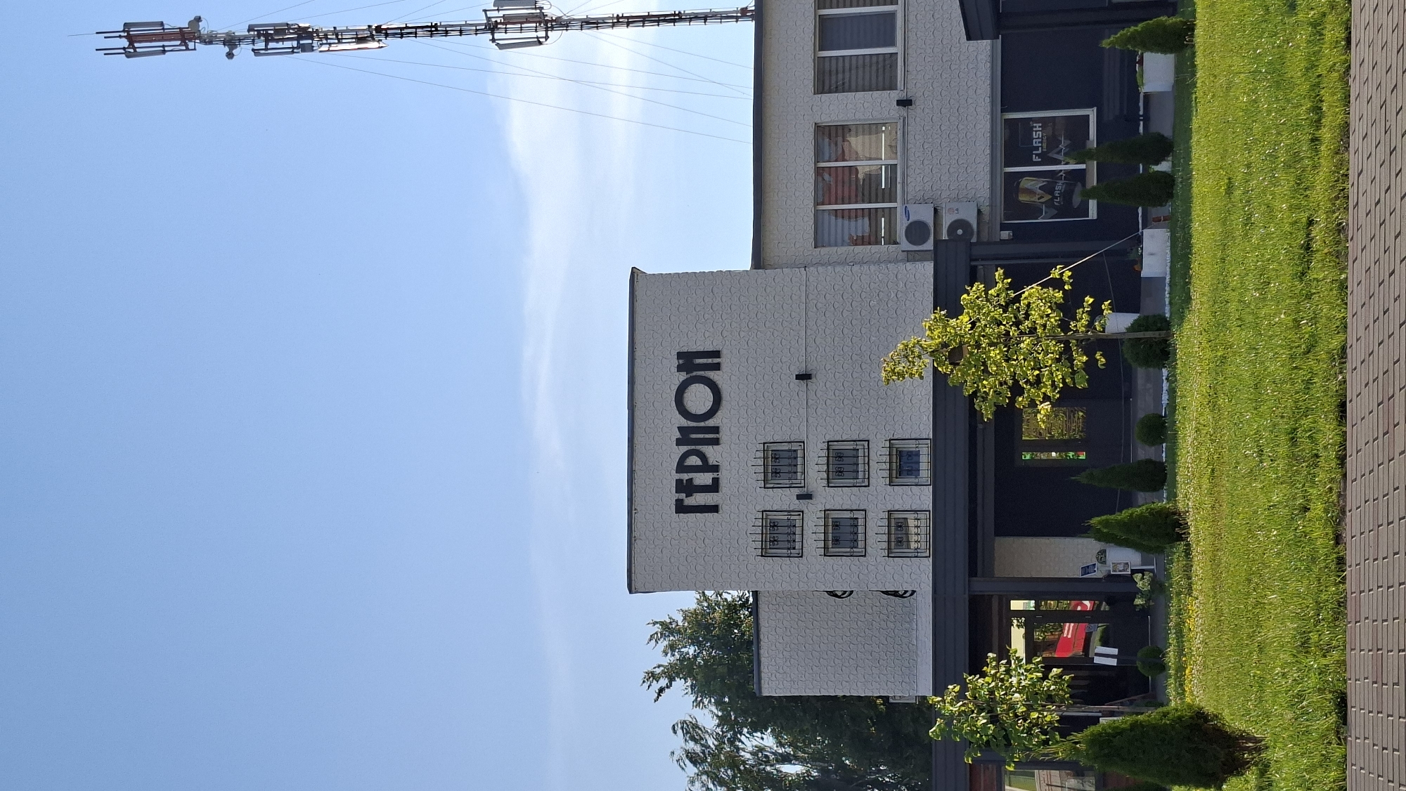 The image depicts a modern building with the Cyrillic text "ГЕРМОН" in large black letters above a row of small, barred windows. The building's facade is textured with a repeating circular pattern and painted a light color. Several windows are visible on the upper floor, some with blinds. An air conditioning unit is mounted on the wall. To the right, a tall cell tower with multiple antennas stands against a clear blue sky. In front of the building, a well-maintained lawn is dotted with small trees and shrubs. A paved walkway is visible in the foreground. The image is taken in daylight, suggesting an outdoor setting in the city of Smalyavichy, Belarus.