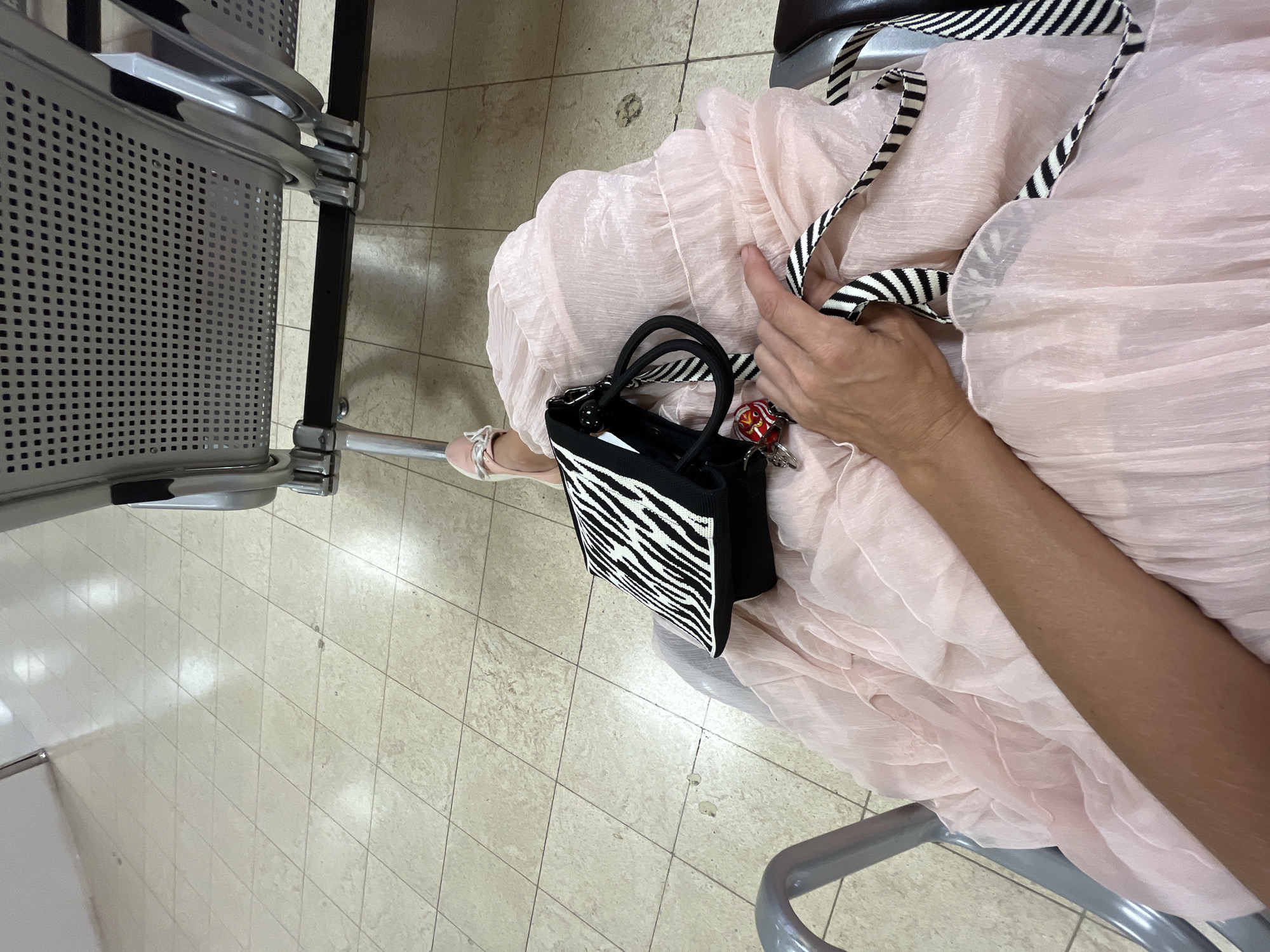 An overhead shot captures a person's hand resting on a light pink, flowing dress. The dress drapes over a person's lap as they sit in a gray metal waiting area chair. A black and white zebra-print handbag is placed on the dress. The bag has two black handles and a black and white striped strap. A small red and gold keychain hangs from the bag. The person's right arm is visible, extending from the lower left of the frame. Their feet, clad in light pink sandals, are visible in the upper left portion of the image, resting on a beige tiled floor. In the background, more gray waiting chairs with perforated seats are visible. The scene appears to be an indoor waiting area, possibly at an airport in Katunayake, Sri Lanka, given the architectural style of the chairs and the tiled floor commonly found in such public spaces.