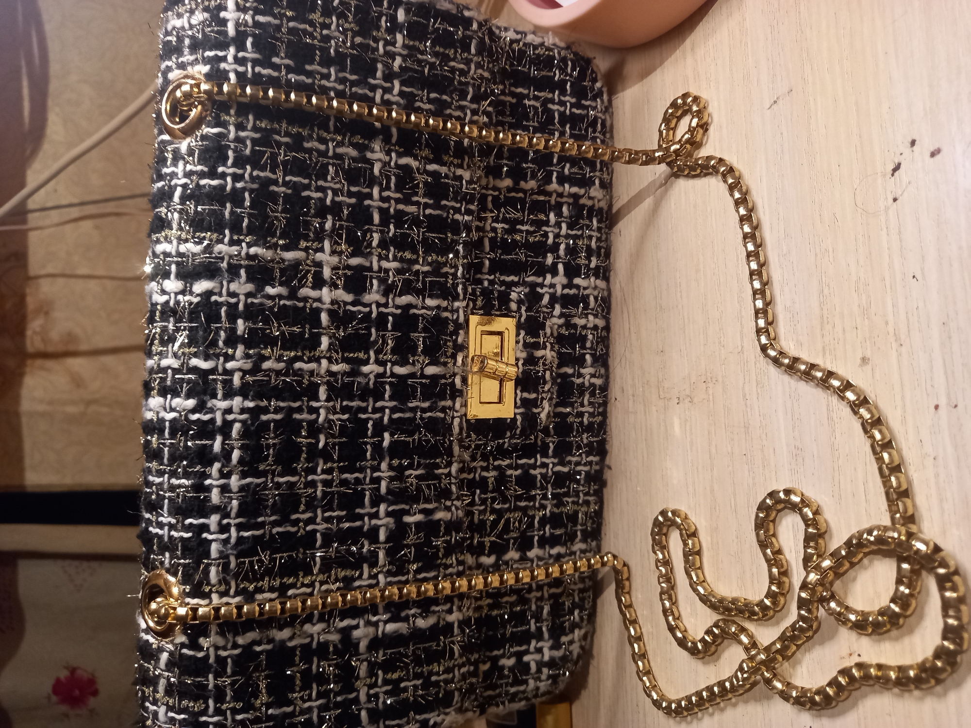 A black and white tweed handbag with gold thread accents and a gold chain strap rests on a light-colored, wooden surface. The handbag features a gold clasp closure on the front. The background consists of a textured wall hanging with a floral pattern. The scene is indoors, likely a room in Sterlibashevo, Russia. No people are visible in the image.