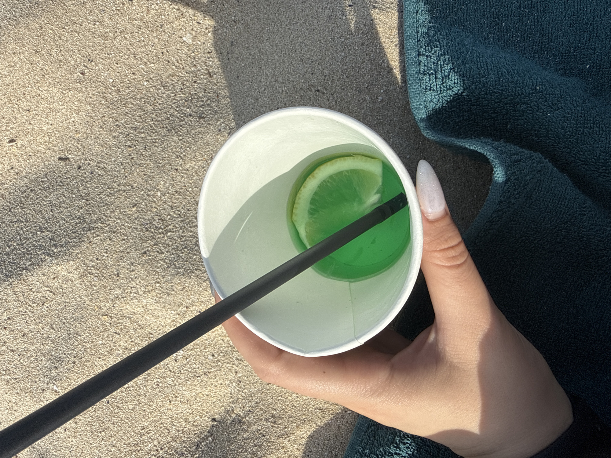 A top-down, high-angle shot captures a person's left hand holding a white paper cup on a sandy surface in an unknown outdoor location. The hand, with light skin tone and a manicured fingernail (white/glitter), grasps the cup from the bottom right. Inside the cup is a vibrant green liquid, a black straw, and a floating slice of lime or lemon. The immediate environment consists primarily of fine-grained, light brown sand, illuminated by strong sunlight casting distinct shadows from the cup and straw. A dark teal-colored terry cloth towel is visible in the lower right corner, beneath the hand and cup. The overall scene suggests a leisurely setting on a beach or sandy area. Location is Unknown, Unknown.