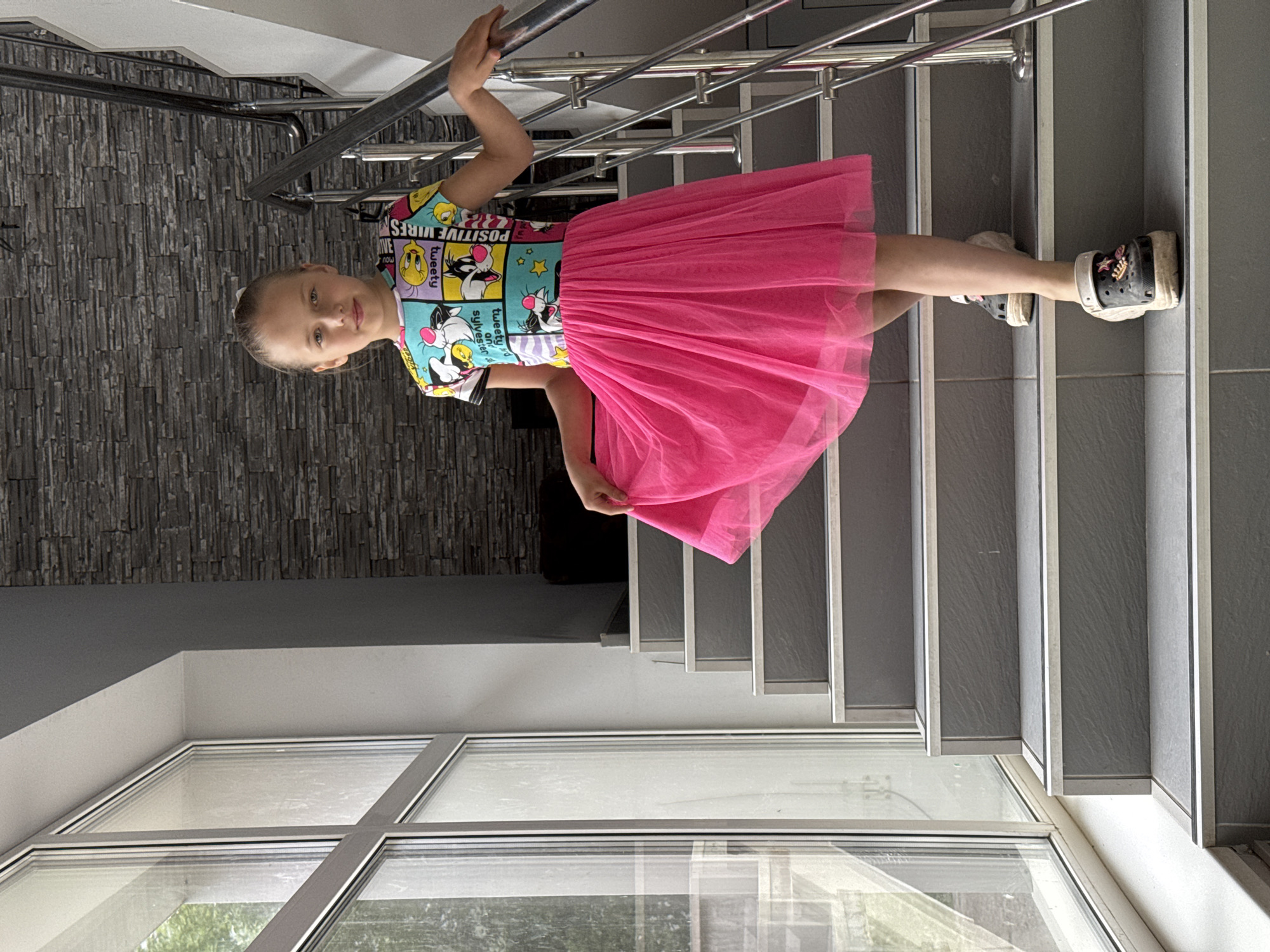 A young girl stands on indoor stairs, holding onto the handrail. She wears a short-sleeved, cartoon-printed shirt and a bright pink tulle skirt. Her footwear consists of black clogs with decorative elements. The stairs are gray with white edges and a metal handrail. The backdrop includes a gray stone wall and a large window showing an outdoor view. The location is Chaikovsky, Russia.