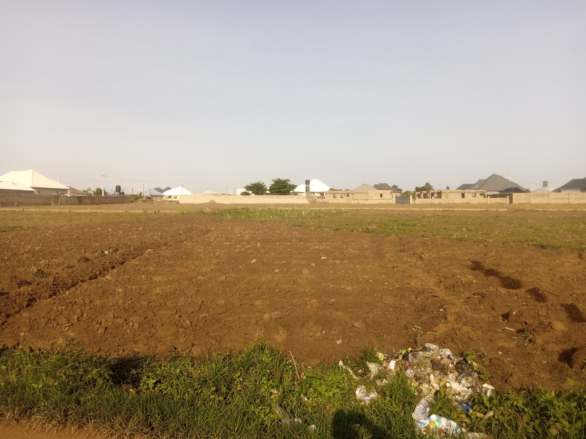 A wide, clear, overcast sky dominates the upper half of the image. Below the sky is a flat, brown field, recently tilled, with rows of soil visible. The field extends from the foreground to the background. In the middle ground, a low, light-colored brick wall stretches across the width of the image, separating the field from a line of buildings. These buildings are mostly single-story structures with modern, peaked roofs, some appearing to be under construction, with exposed brickwork and concrete foundations. Among the buildings, there are a few trees. In the foreground, along the bottom edge of the image, is a strip of green grass and weeds, with a pile of assorted trash, including plastic bags and bottles, situated on the right side. The overall scene depicts a developing or partially developed residential area on the outskirts of a city.