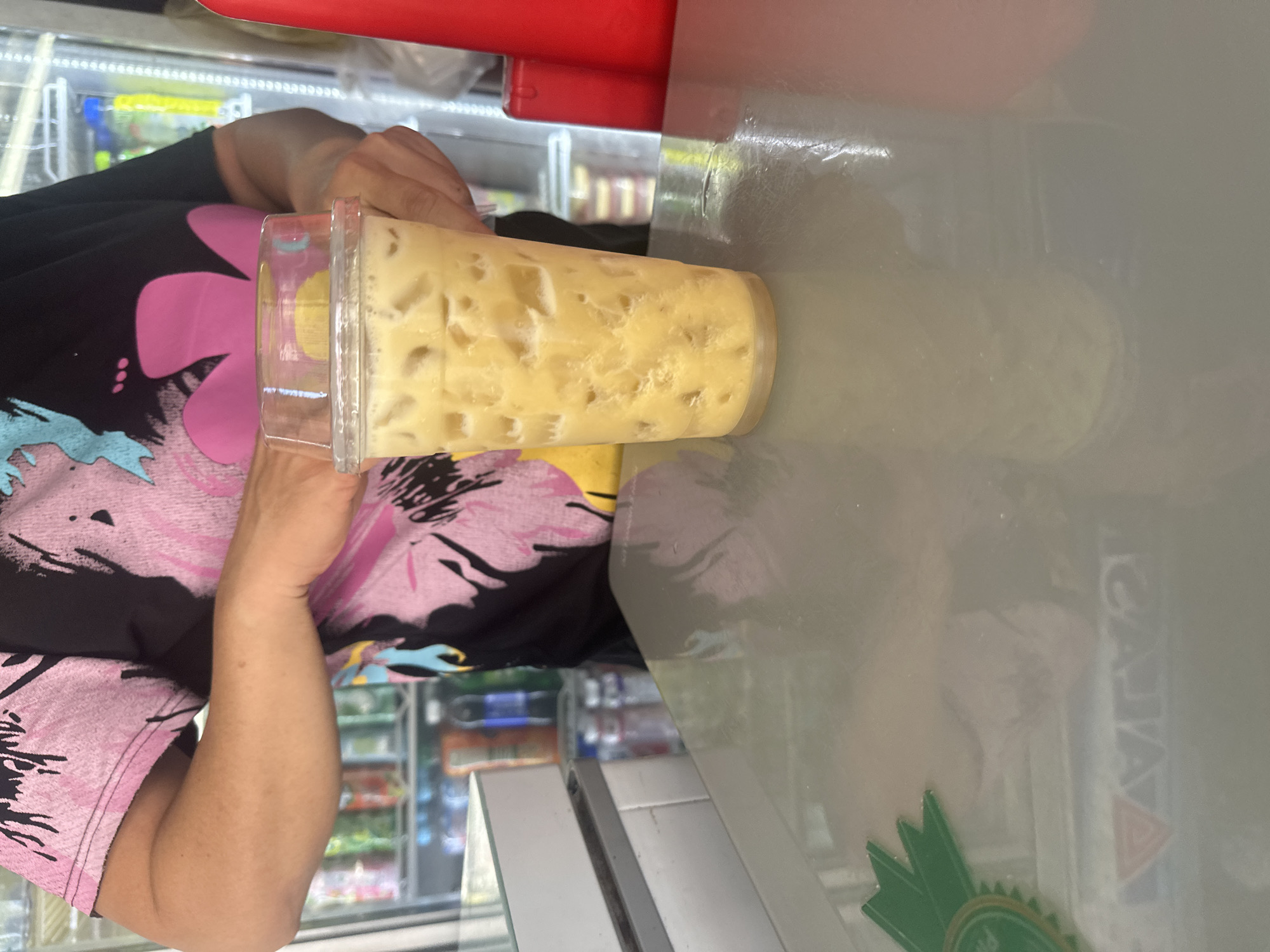 A person is holding a yellow drink with ice in a clear plastic cup. The background shows the inside of a shop with shelves stocked with various items. The shop is possibly located in Bac Nha Trang Ward, Vietnam. The person is wearing a shirt with a floral design. The drink is placed on a surface, likely a counter, with a green logo visible on it. A refrigerator with glass doors is seen in the background, filled with products.