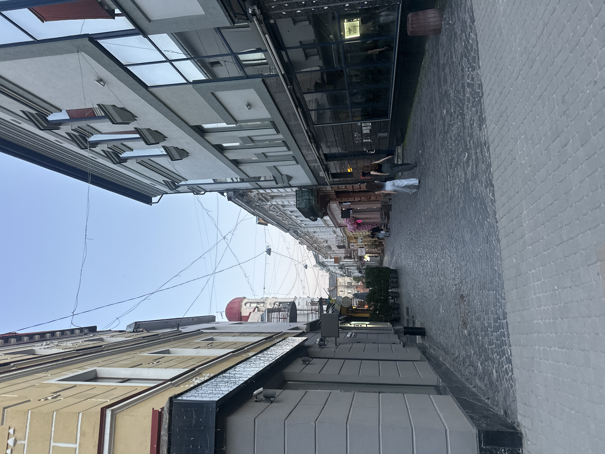 The image depicts a street scene in Ternopil, Ukraine, likely during twilight hours. The perspective is from the street level, looking down a cobblestone alley lined with buildings. On the left, a pale yellow building with white trim has multiple windows and what appears to be a storefront at ground level, partially obscured by the angle. On the right, a modern building with large, reflective glass windows dominates the view.
Further down the street, other buildings with a mix of architectural styles are visible, including one with a red dome-like structure and balconies. The street is paved with cobblestones and some areas are lined with grey paving stones. Potted plants are visible outside some establishments.
Two individuals, likely women, are walking away from the camera down the street on the right side. They are dressed in casual clothing. The sky above is a clear, pale blue, indicating either early morning or late evening. Overhead, numerous electrical wires are strung across the street, crisscrossing the sky. The overall impression is of a quiet, urban street in the late day.