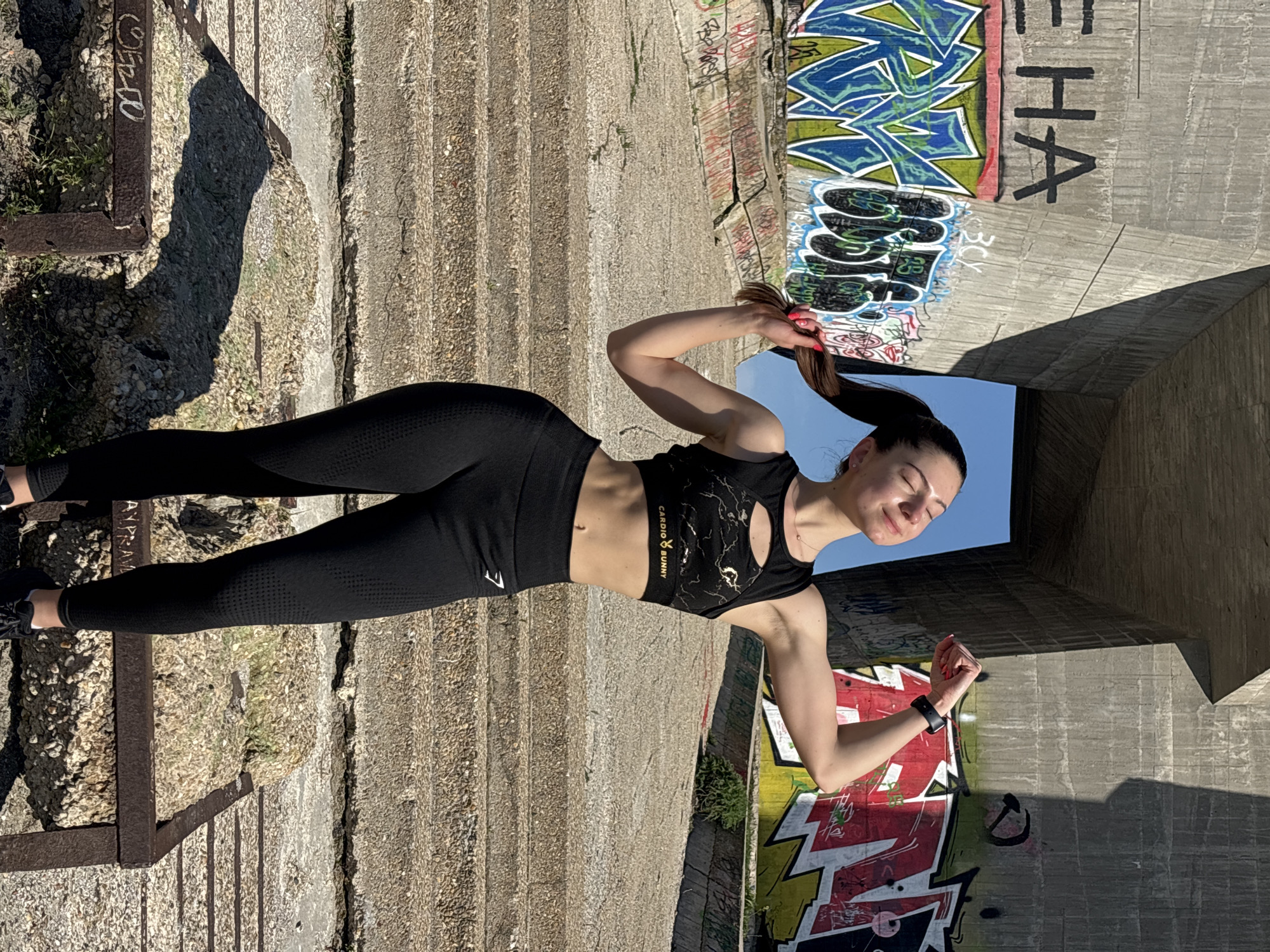 An athletic young woman poses outdoors in a sunlit, urban environment. She stands with her body slightly turned, holding her high ponytail with her right hand, while her left arm is bent. Her eyes are closed, and she has a relaxed, content expression, basking in the sunlight. She is wearing a black athletic ensemble, including a sports bra with cutouts and a gold-speckled or marble-patterned design, and form-fitting black high-waisted leggings with a textured pattern. A black smartwatch is visible on her left wrist.
The setting appears to be an industrial or abandoned concrete structure, possibly an underpass or a forgotten outdoor area. The ground is rough, uneven concrete or gravel, with sparse weeds growing through cracks. To the left, a rusty metal frame is partially visible. The background consists of large, textured concrete walls, heavily adorned with colorful graffiti. Prominent blue and green abstract designs are visible, along with black and white tags. A large black "HA" is painted vertically on a concrete column to the right. The woman's shadow is cast long on the ground, indicating bright sunshine, likely morning or late afternoon. Above, a clear blue sky is visible through a gap in the concrete structure.
Visible text includes "CARDIO BUNNY" and a ribbon logo on her sports bra. A white logo, possibly "GYMSHARK" or a similar brand, is on her left thigh. Graffiti on the walls includes "3CY", other illegible tags, and what might be "AYPRAM" on a lower wall or ground section.