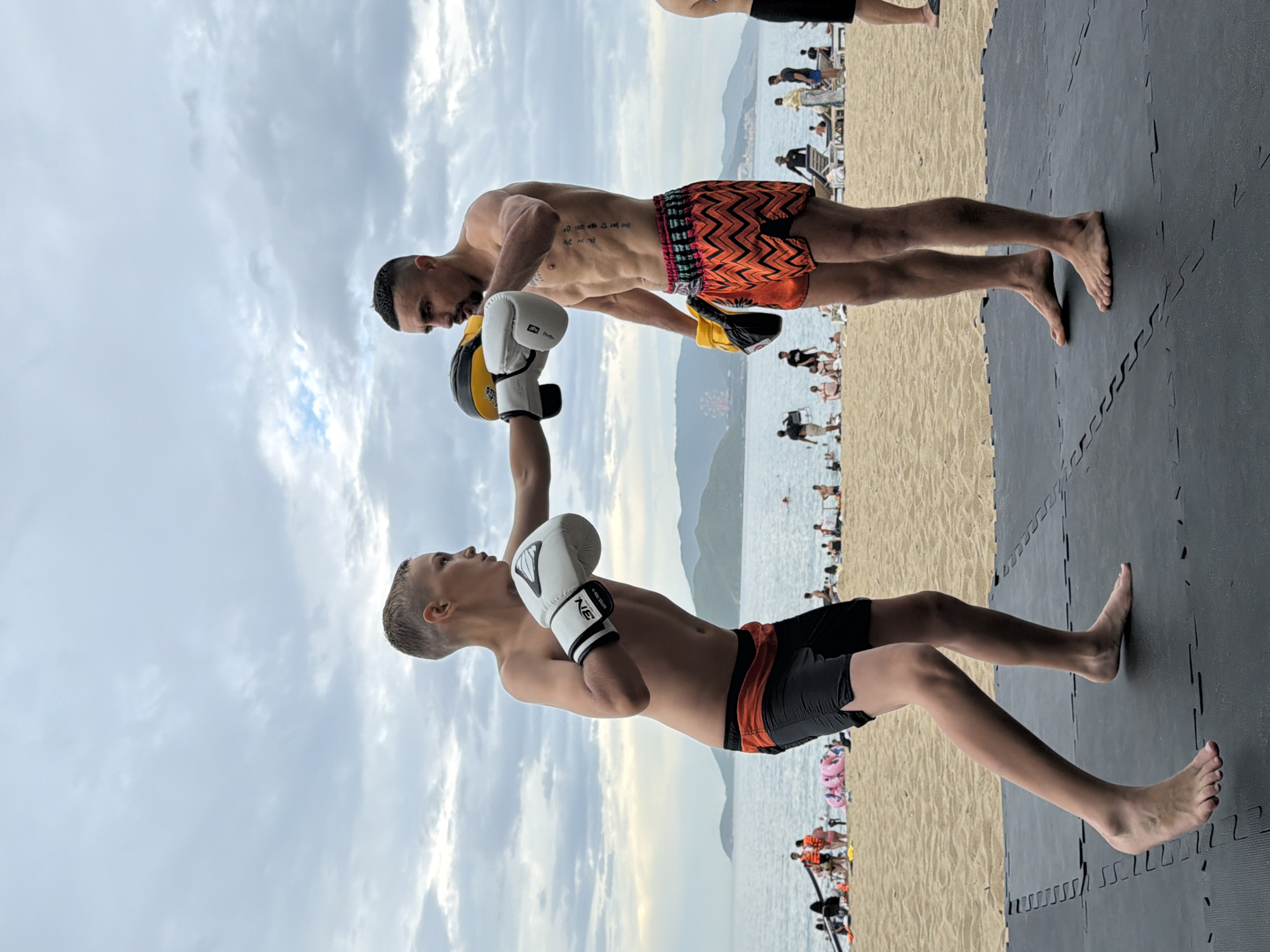 The image shows two males practicing martial arts on a beach in Nha Trang, Vietnam. The older male, with a muscular physique and tribal tattoos on his torso, is wearing orange and black patterned shorts. He is holding a yellow and black padded glove, likely for training. The younger male, who appears to be a boy, is wearing black shorts and white boxing gloves. He is in a defensive stance, facing the older male.
The scene is set on a beach with sand leading to the ocean in the background. The sky is cloudy, suggesting it might be overcast or nearing sunset. In the distance, there are mountains and the ocean, with a few boats or buoys visible. There are also other people on the beach, some appearing to be sunbathing or engaging in recreational activities. The practice is taking place on a black mat, which is laid out on the sand, providing a stable surface for their training.
The overall mood appears focused and disciplined, with both individuals concentrating on their training. The lighting suggests it is either early morning or late afternoon. The presence of the ocean, mountains, and numerous people on the beach strongly indicates a coastal location. No visible text is discernible in the image, other than the brand name "NIE" on the younger male's boxing glove.