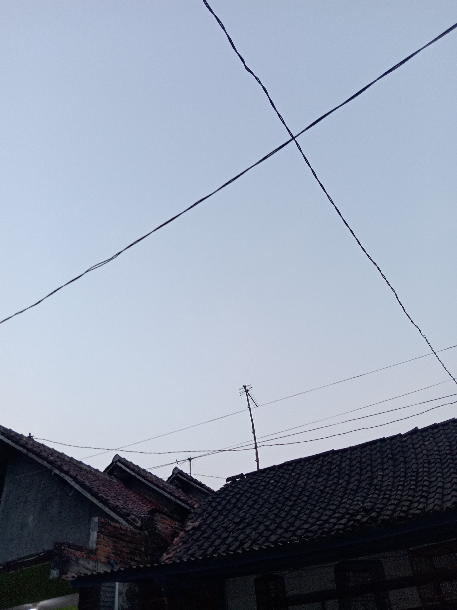 The image shows a view of rooftops in Jumput, Indonesia, against a pale, overcast sky. Two thick, black wires cross each other in the sky, creating an "X" shape. Below the wires, several dark-tiled rooftops are visible, with varying pitches and designs. Some of the rooftops appear weathered. A metal antenna stands atop one of the houses. The lower part of the image shows the dark facades of some buildings, hinting at their presence beneath the roofs. No people are visible in the frame. The scene suggests an ordinary residential area in the city.