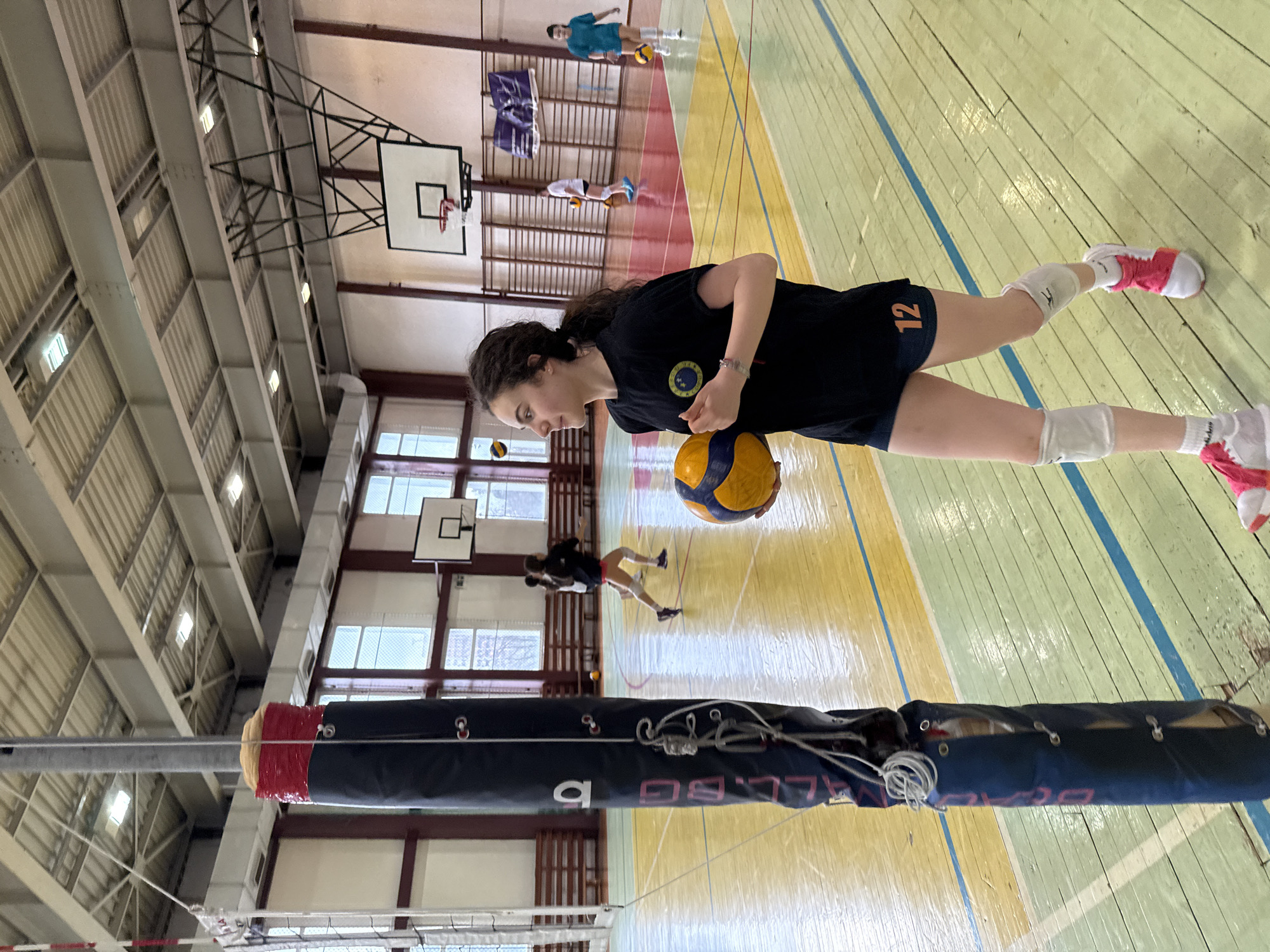 A young woman in a black t-shirt and shorts, knee pads, and pink-accented sneakers stands on a multi-colored gymnasium floor with a volleyball in her hands. The number "12" is visible on her shorts. A volleyball net with a padded pole is in the foreground. In the background, other individuals can be seen on the court with basketball hoops visible in the background. The scene is located in the city of Sofia, Bulgaria.