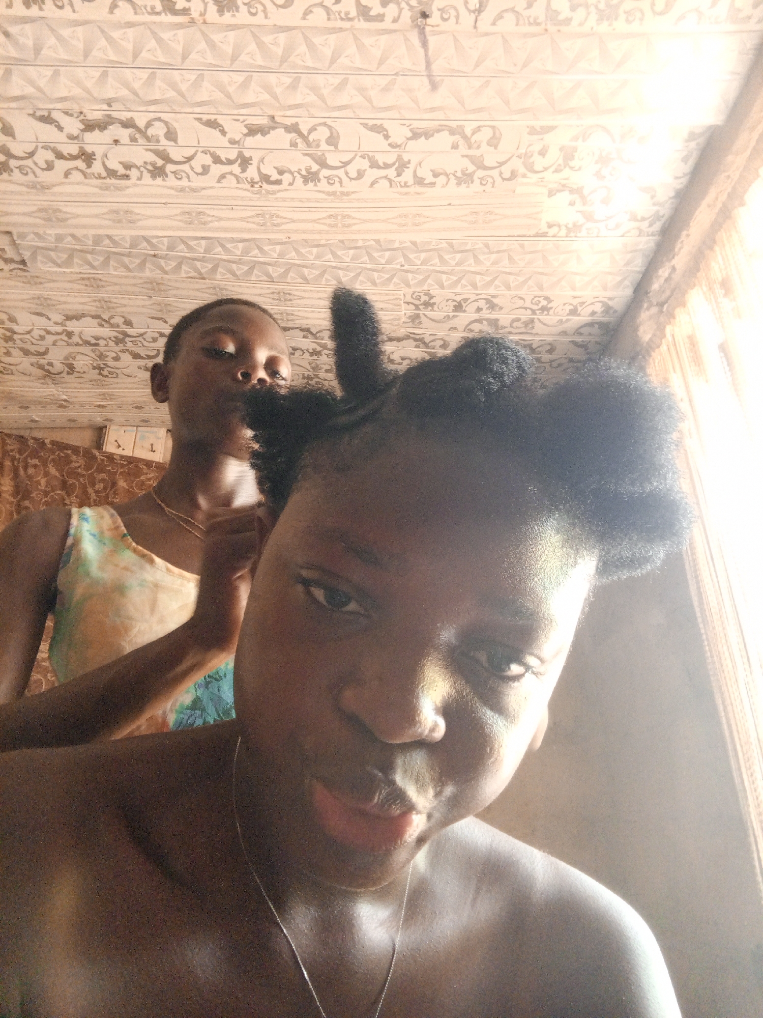 The image depicts two Black females indoors. The individual in the foreground appears to be taking a selfie, focusing on her face and upper torso. She is wearing a silver necklace. The second female stands slightly behind her, wearing a multi-colored patterned top and a gold necklace. Her hands are raised near the first female's head, potentially styling her hair. The background reveals an interior with textured ceiling tiles, a window with sheer curtains, and a glimpse of a patterned wall. The location is likely a residential interior in Uyo, Nigeria.