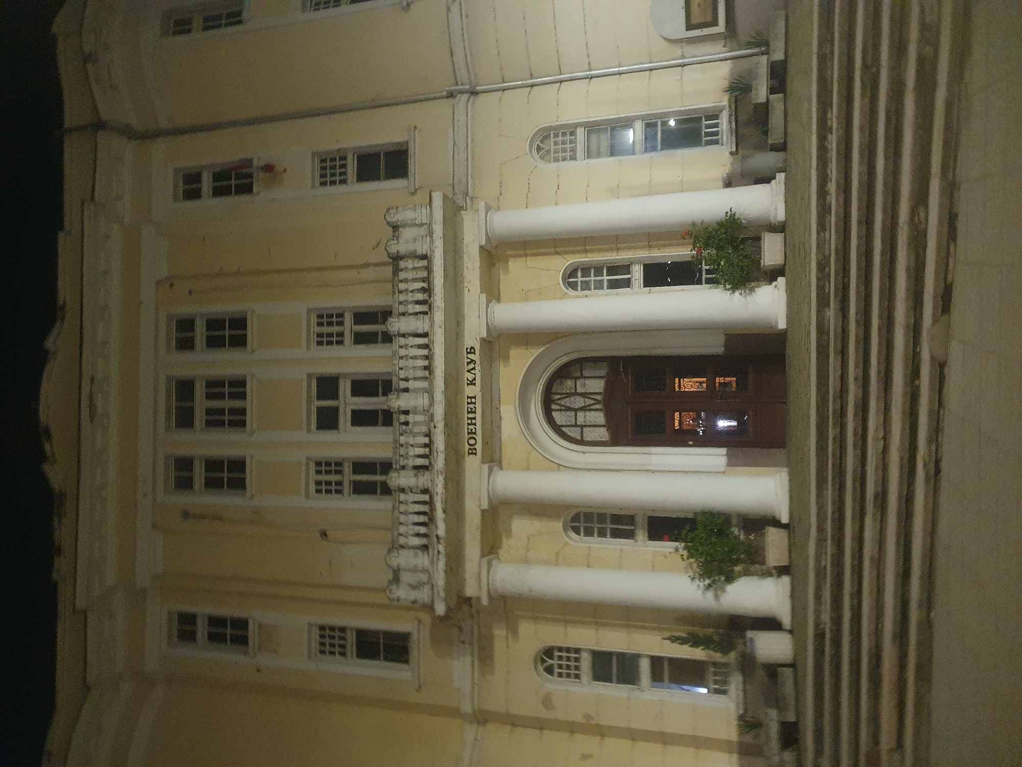 The image shows the facade of the Plovdiv Military Club in Bulgaria at night. The building is a yellow, multi-story structure with classical architectural elements, including white columns and arched windows. A sign above the entrance reads "ВОЕНЕН КЛУБ" (Military Club). There are plants in decorative planters on the balcony. The image is somewhat blurry and taken at an angle.