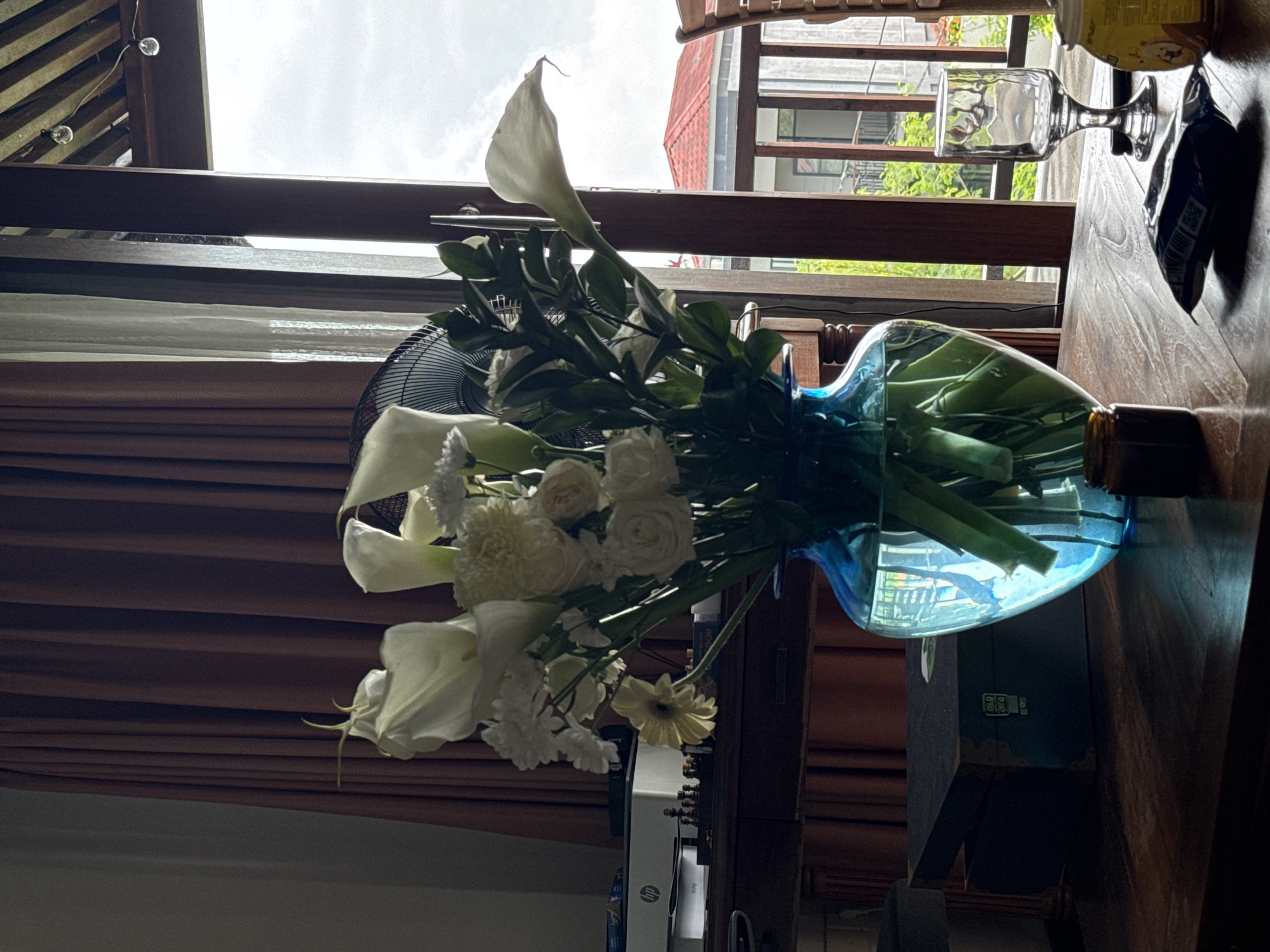 The image, potentially taken indoors in Kuta Selatan, Indonesia, shows a bouquet of white flowers, including calla lilies and roses, in a blue glass vase. The vase is situated on a dark wooden table. A standing fan is behind the bouquet. A drinking glass and a bottle are also present on the table. Outside the window, buildings and foliage are visible. Brown curtains hang near the window.