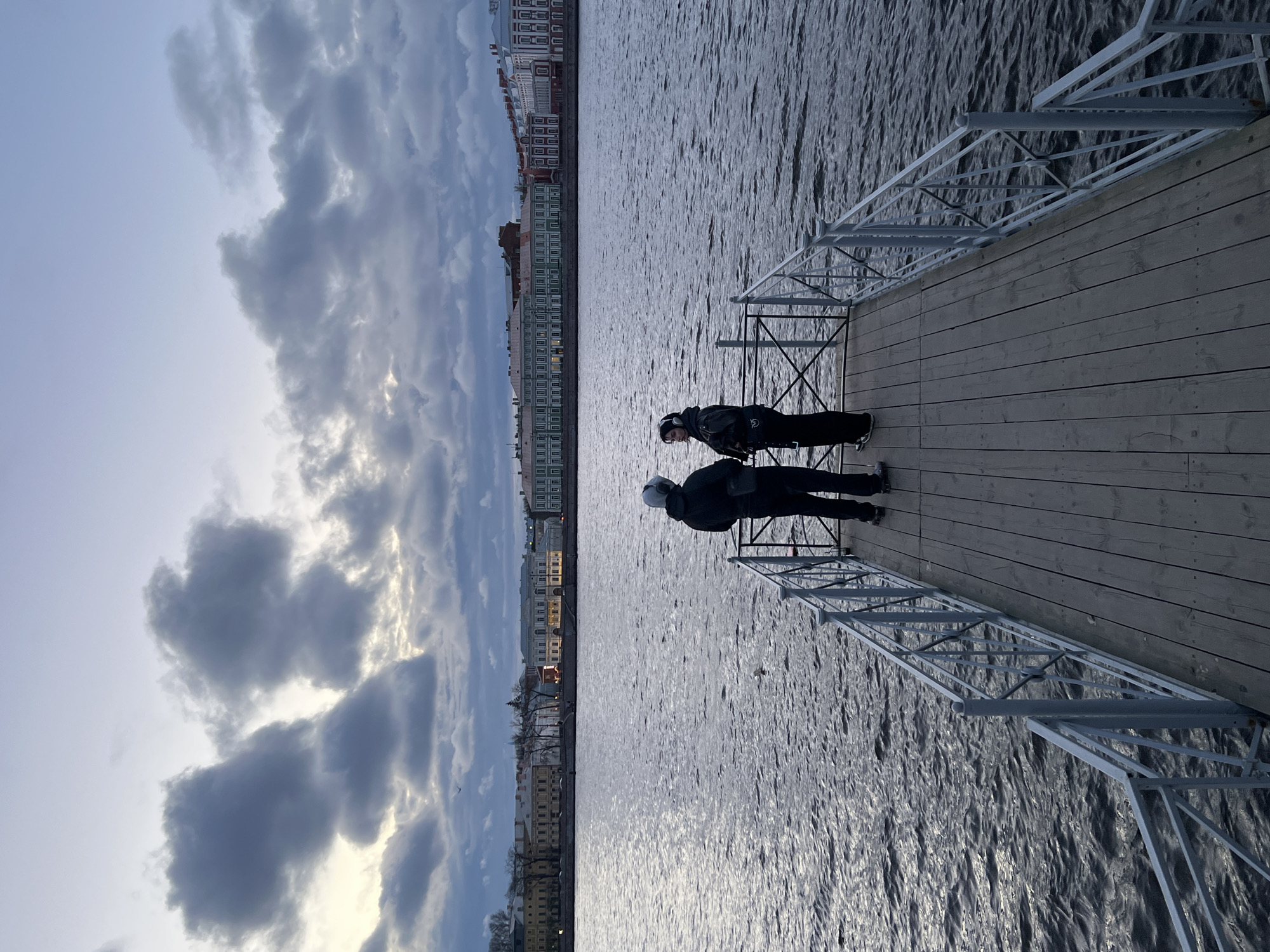 Two people stand on a wooden pier with metal railings, overlooking a body of water. The water reflects the city buildings across the way, characteristic of the architecture in Saint Petersburg, Russia. The sky is overcast with dense clouds.