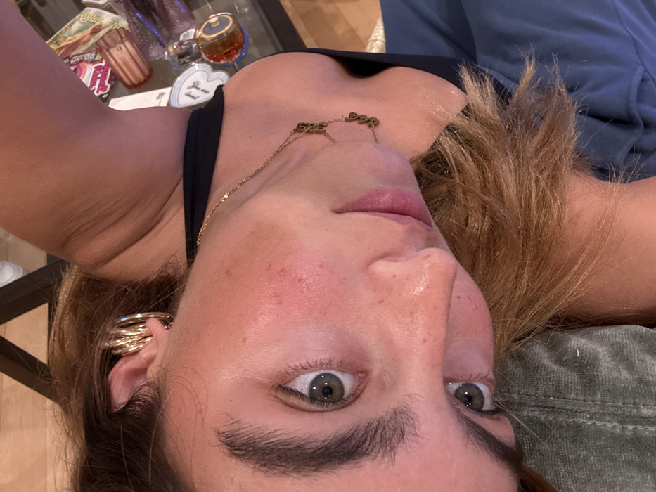The image displays a close-up, high-angle view of a female subject, oriented approximately 65 degrees counter-clockwise from a standard upright perspective. The scene is indoors, in what appears to be a residential setting within Los Angeles, United States of America.
The subject is a young adult female with light brown hair worn loosely around her shoulders. She has fair skin, with visible freckles and patches of redness on her right cheek and chin. Her eyes are light-colored, possibly green or hazel, and she is looking directly at the camera. Her eyebrows are dark and full. She wears a black top, possibly a tank top or camisole. On her right ear, multiple gold-colored ear cuffs or rings are visible on the helix cartilage. A delicate gold-colored chain necklace with small, dark, letter-like charms, possibly spelling "$$$", hangs around her neck, resting on her collarbone area. Her lips are slightly parted, revealing a light pink color.
In the blurred background, to the upper right of the subject, a clear glass-top table is visible. Several items rest on the table, including: a dark, possibly amber-colored perfume bottle with a metallic cap; a white heart-shaped object with dark markings, possibly "FF"; another small, clear bottle with a heart-shaped stopper; and a rectangular object, possibly a decorative box or wallet, featuring a colorful illustration. Below the subject's left shoulder, a section of dark blue fabric is visible, possibly clothing or upholstery. Below her head, a textured olive-green fabric, possibly a pillow or blanket, is present. The lighting is consistent with indoor ambient light. No discernible interactions with other individuals are depicted.