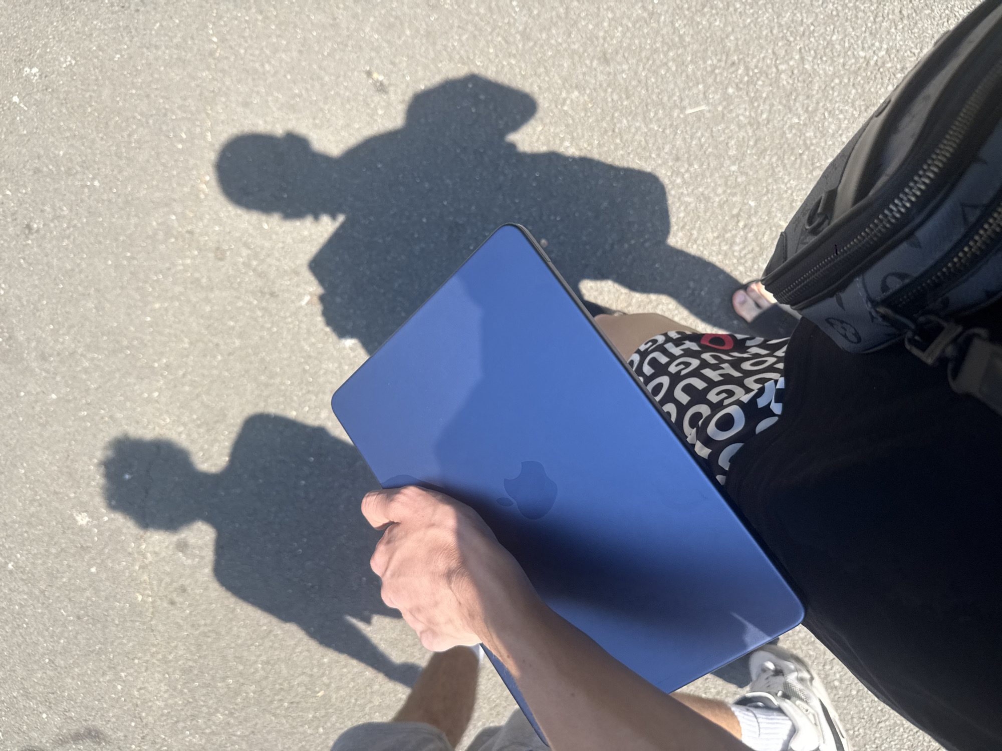 A person's hand holds a blue laptop with an Apple logo visible on its surface. The laptop is positioned on a paved surface, casting a shadow. In the background, another, larger shadow of a person is visible on the ground. The person holding the laptop is wearing a black shirt with a white patterned design and white sneakers. They are also carrying a black bag with a patterned design. The scene suggests an outdoor setting, possibly during daytime due to the shadows, and the lighting implies it might be a sunny day. The overall tone appears casual and practical, with the person seemingly on the go with their laptop. The location is identified as Burgas, Bulgaria. No visible text is present in the image.