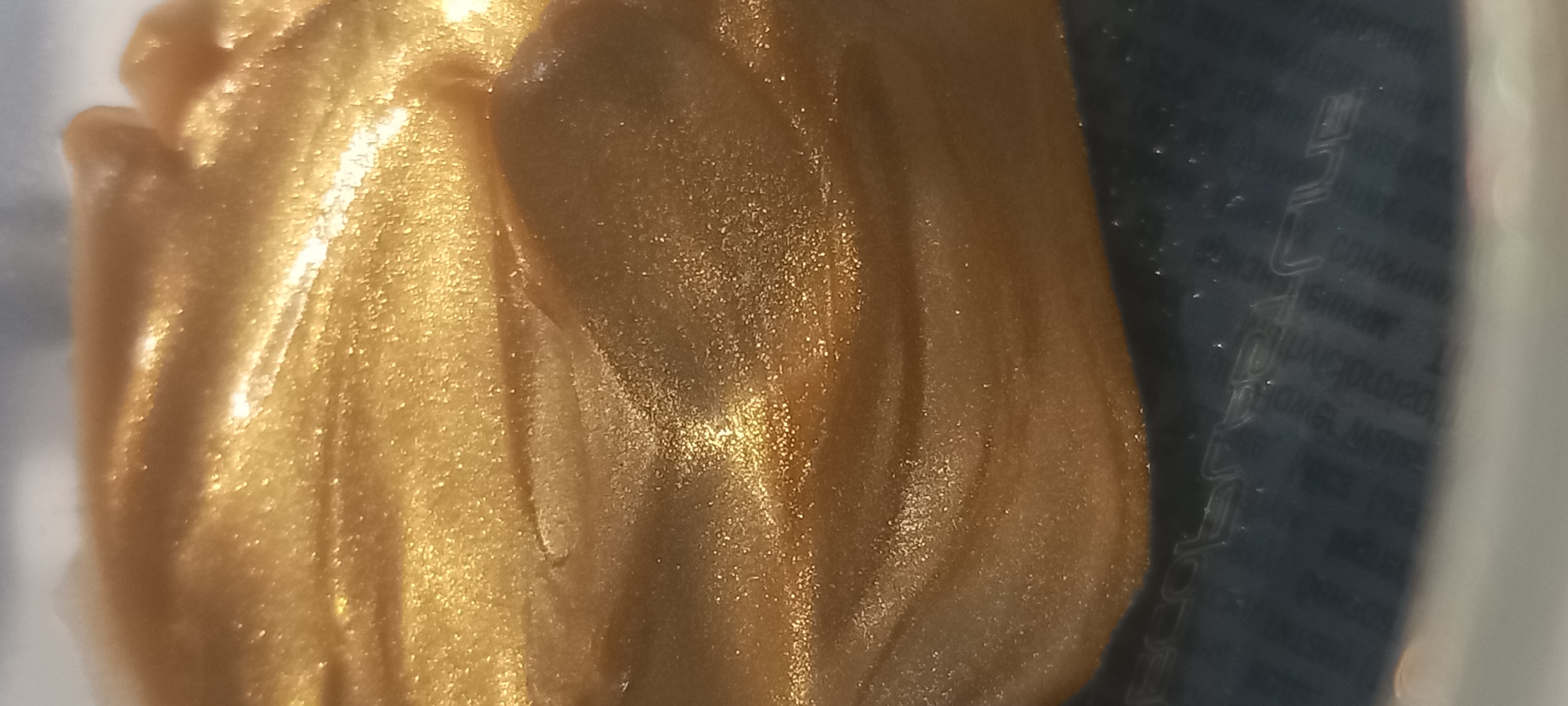 The image is a close-up, abstract shot of a swirl of golden, shimmering lotion or cream. The texture is smooth with subtle undulations, and it reflects light, creating highlights and shadows that give it a luminous quality. Tiny sparkles are visible within the substance, suggesting a cosmetic product with glitter. In the lower portion of the image, a dark, reflective surface is visible, on which some faint embossed text can be discerned. The text appears to be a product label, with letters like "ECOLE" and "PLUG" partially visible. The overall impression is one of luxury and radiance, likely a cosmetic product. The image does not contain any identifiable landmarks or elements that would allow for its placement in Kryvyi Rih, Ukraine, or any other specific city.