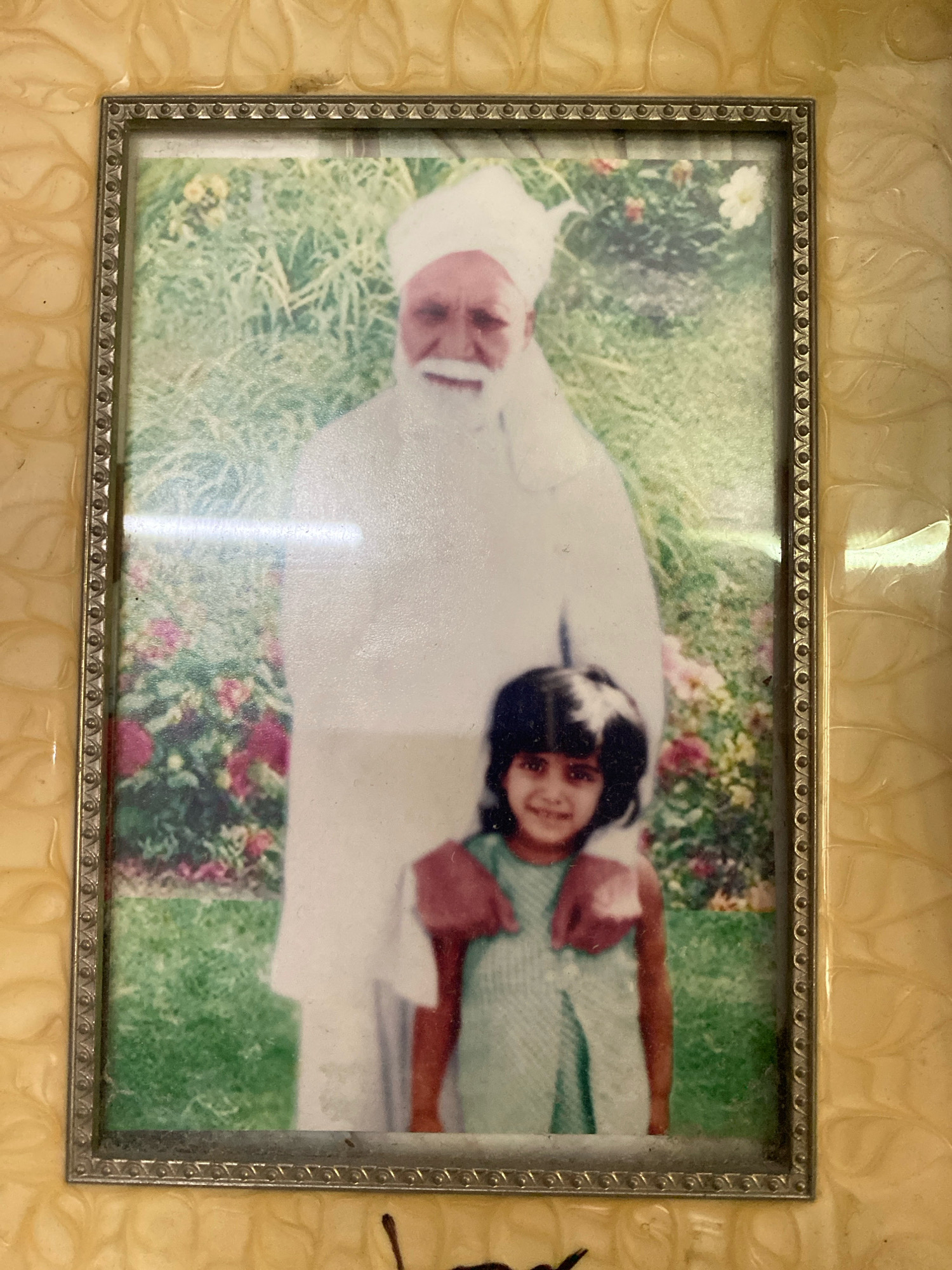 A framed photograph depicts an elderly man and a young girl standing outdoors. The man, positioned behind the girl, wears a white turban and a white garment. He has a beard and is smiling. The young girl, in the foreground, wears a light blue and white striped dress. Her hands are on the man's shoulders, and she is also smiling. The background features green grass and colorful flowers, suggesting a garden setting. The photograph is enclosed in a decorative frame with an embossed, swirling pattern. The image is a photograph of a photograph, with some glare and minor imperfections visible. The scene is set in the city of Keer, Pakistan.