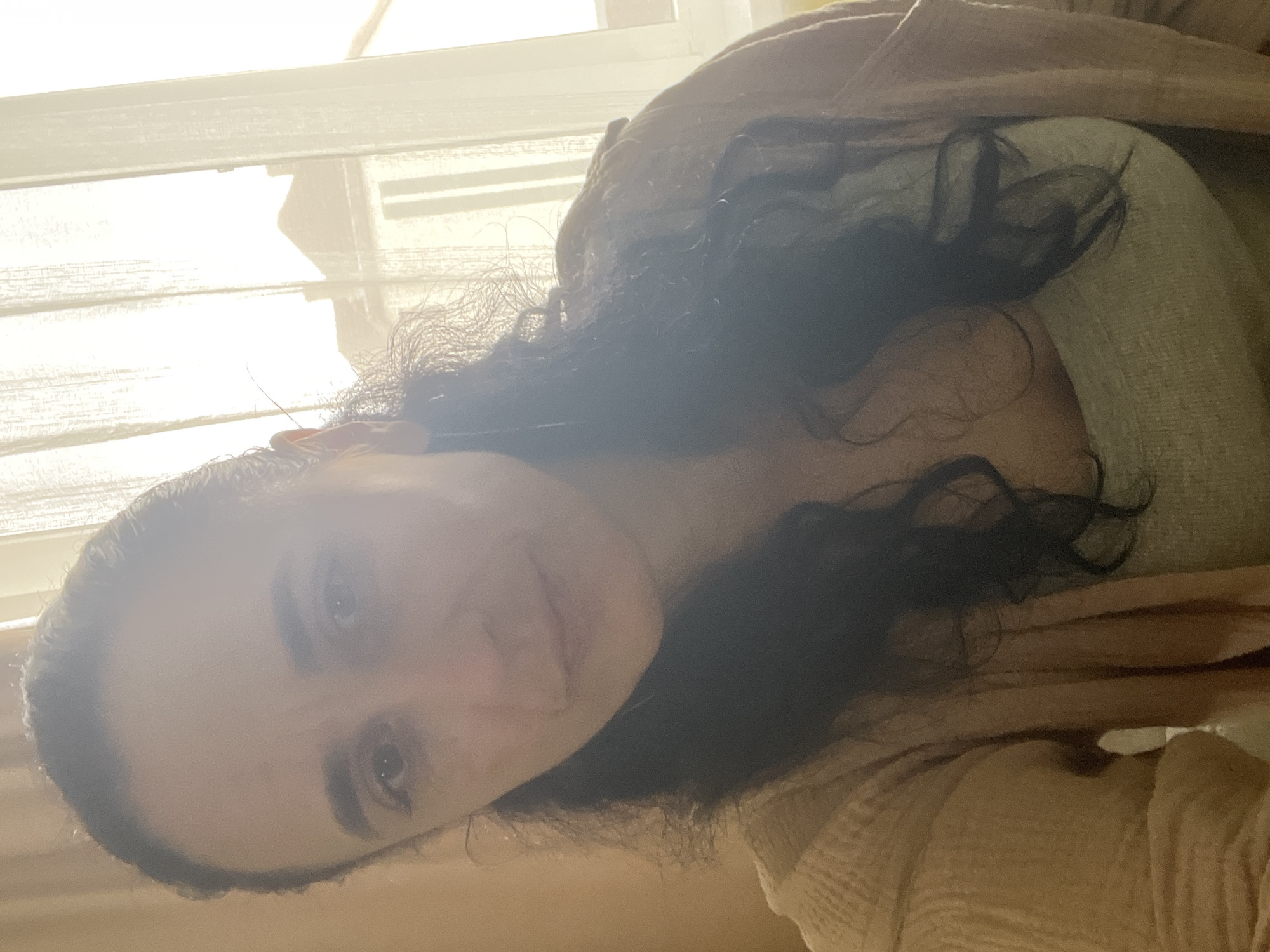 This portrait, rotated 90 degrees counter-clockwise, depicts a person, likely a woman, positioned indoors in Onokivtsi, Ukraine. The subject has long, dark, curly hair and is looking directly at the camera with a neutral to slightly smiling expression. She is wearing a light-colored, possibly beige or pale orange, textured garment over a lighter inner shirt.
The background features a brightly lit window with blinds or sheer curtains, indicating it is daytime. The strong light source causes significant glare and overexposure in the upper and left portions of the image, obscuring some detail and creating a soft halo effect around the person's head. Visible details include the texture of her clothing and the individual strands of her dark, wavy hair. No specific activities other than posing for the photo are evident, and no text is visible in the frame. The image conveys a bright, casual setting.