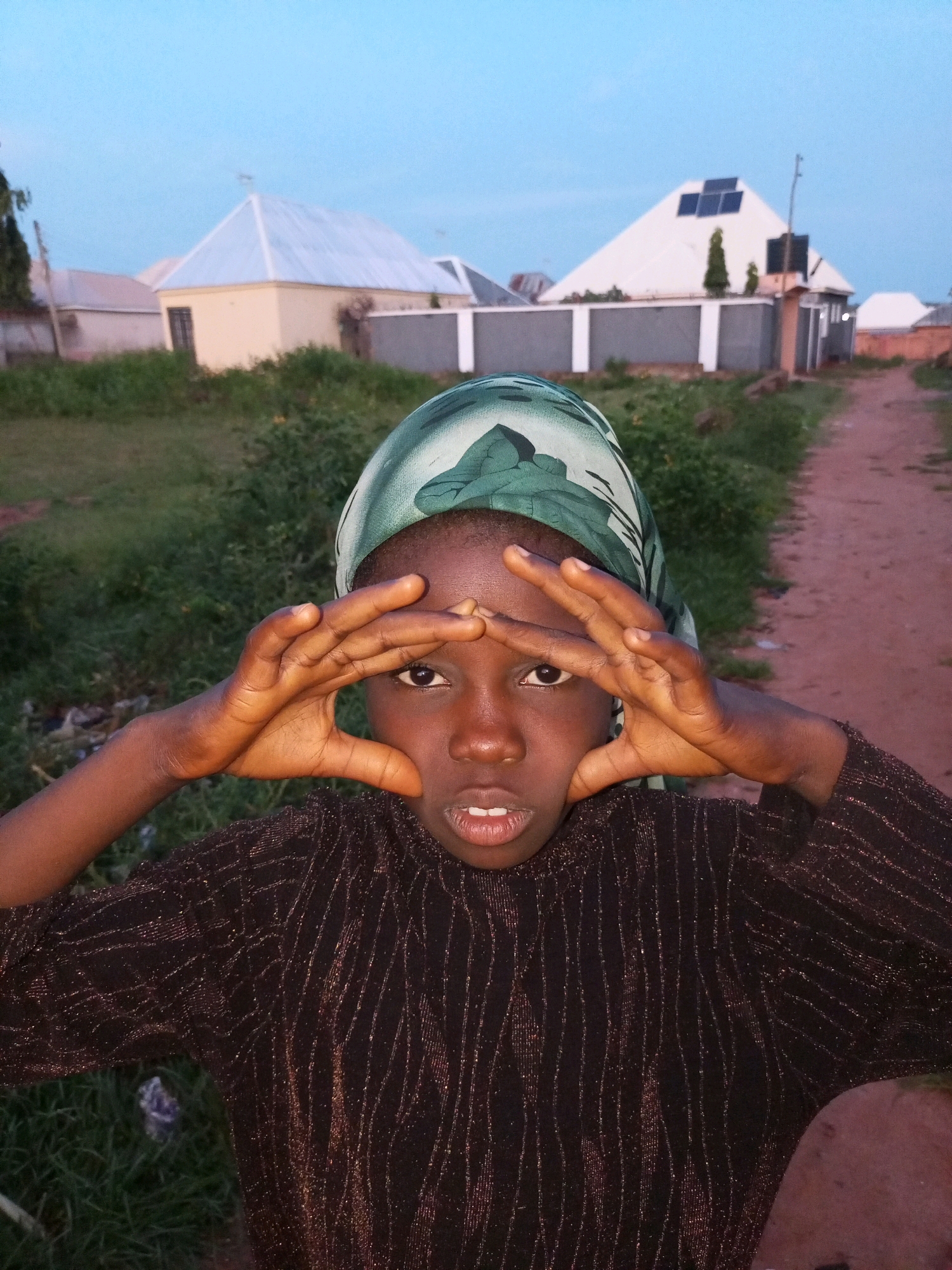 A young girl, wearing a green patterned headscarf and a dark, shimmering long-sleeved shirt, is pictured in an outdoor setting. She is looking directly at the camera, with her hands raised to frame her eyes in a playful gesture. Her mouth is slightly open, revealing her teeth. The background features several buildings with distinct roof structures, one of which has solar panels. A dirt path runs to the right of the frame, and there is green grass and vegetation in the foreground and mid-ground. The sky is a pale blue, indicating it is either early morning or late evening. The location is identified as Tempure, Nigeria.