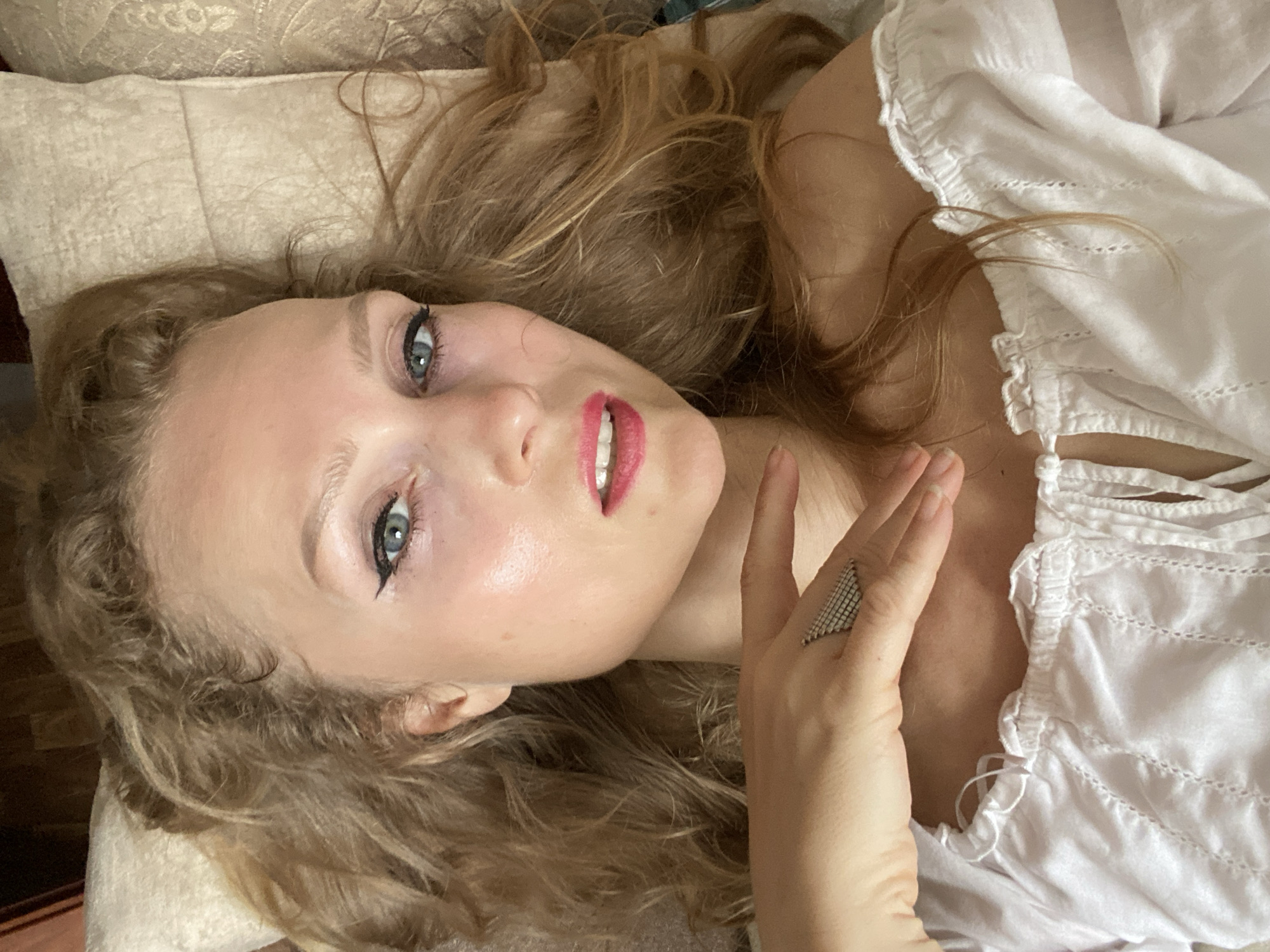 A close-up, overhead shot shows a young woman with long, wavy blonde hair lying on her back on a beige, textured surface. Her eyes are open and looking towards the camera. She has on bold black eyeliner and bright pink lipstick. Her skin appears lightly powdered and has a rosy sheen on her cheeks. Her right hand is raised, fingers splayed, with a geometric, silver ring on her index finger. She is wearing a white, off-the-shoulder top with ruffled edges and decorative stitching. The background consists of what appears to be a headrest or pillow. The lighting is bright and casts subtle shadows. There is no indication of the city of Lida, Belarus in the image.