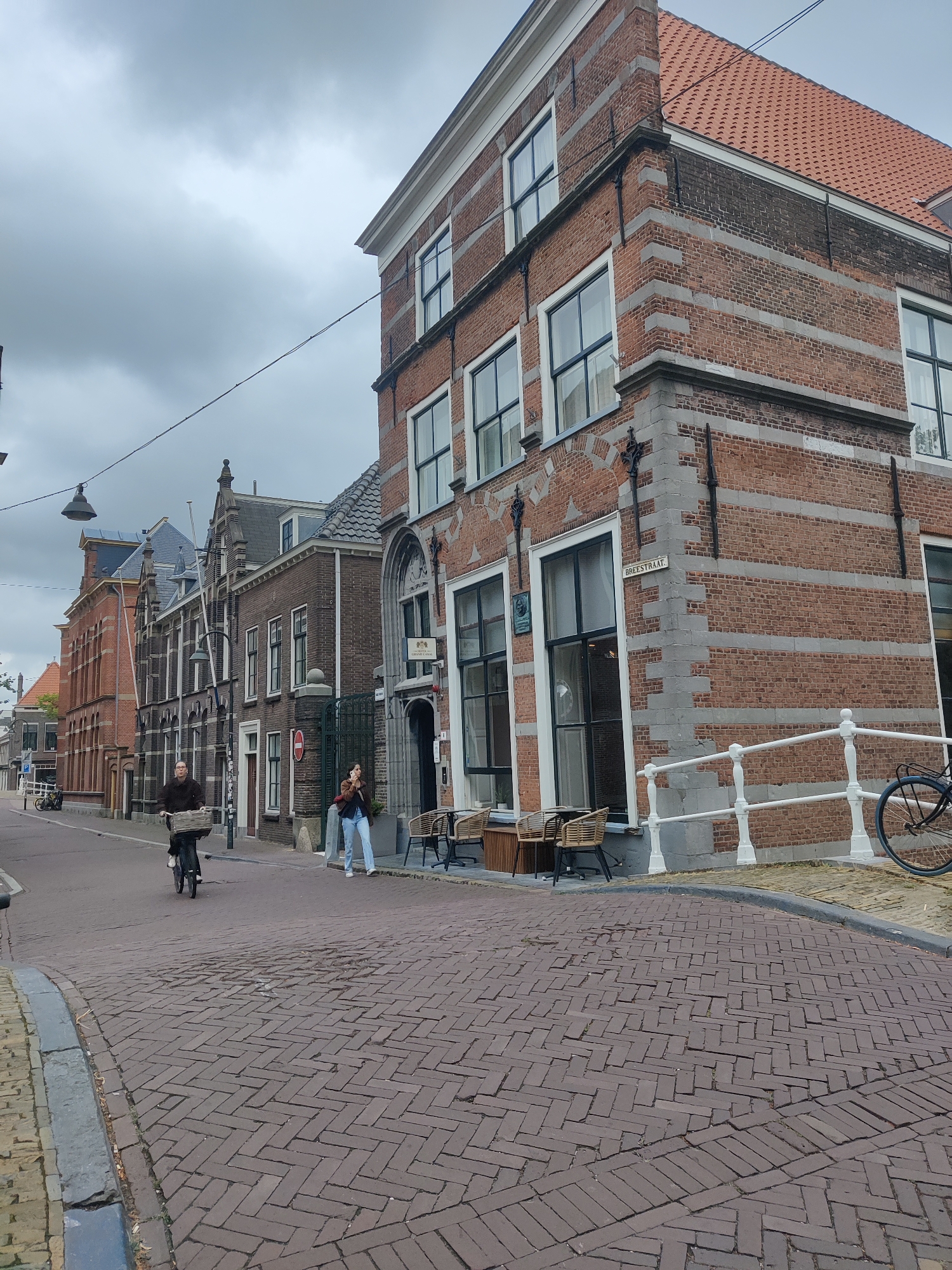 The image captures a street scene in Delft, Netherlands, on an overcast day. The perspective is from street level, looking up at buildings and a section of the street.
In the foreground, the street is paved with red bricks laid in a herringbone pattern. A curb runs along the left side of the street, transitioning to a yellow brick sidewalk. To the right, a white railing with decorative posts separates the sidewalk from a body of water, where a bicycle is parked and leaning against the railing.
Dominating the right side of the frame is a tall, brick building with a red tile roof. The building features multiple stories with large, dark-framed windows. The brickwork is characterized by alternating horizontal bands of lighter and darker bricks, with decorative stone quoins at the corners. A sign above a doorway reads "BREEESTRAAT". Outside this building, a small outdoor seating area with woven chairs and small tables is visible.
Further down the street, several other buildings line the left side. These buildings are also constructed of brick, with varying architectural styles and rooflines. Some have arched windows and decorative stonework. Streetlights are visible, including one with a distinctive lantern-style fixture.
Two individuals are present in the scene. A person is riding a bicycle towards the viewer on the brick-paved street, with a basket on the handlebars. Another person is standing on the sidewalk near the building with the "BREEESTRAAT" sign, holding a phone to their ear.
The sky is heavily overcast with grey clouds, suggesting a dull or potentially rainy day. The overall impression is one of a historic European street with traditional architecture.