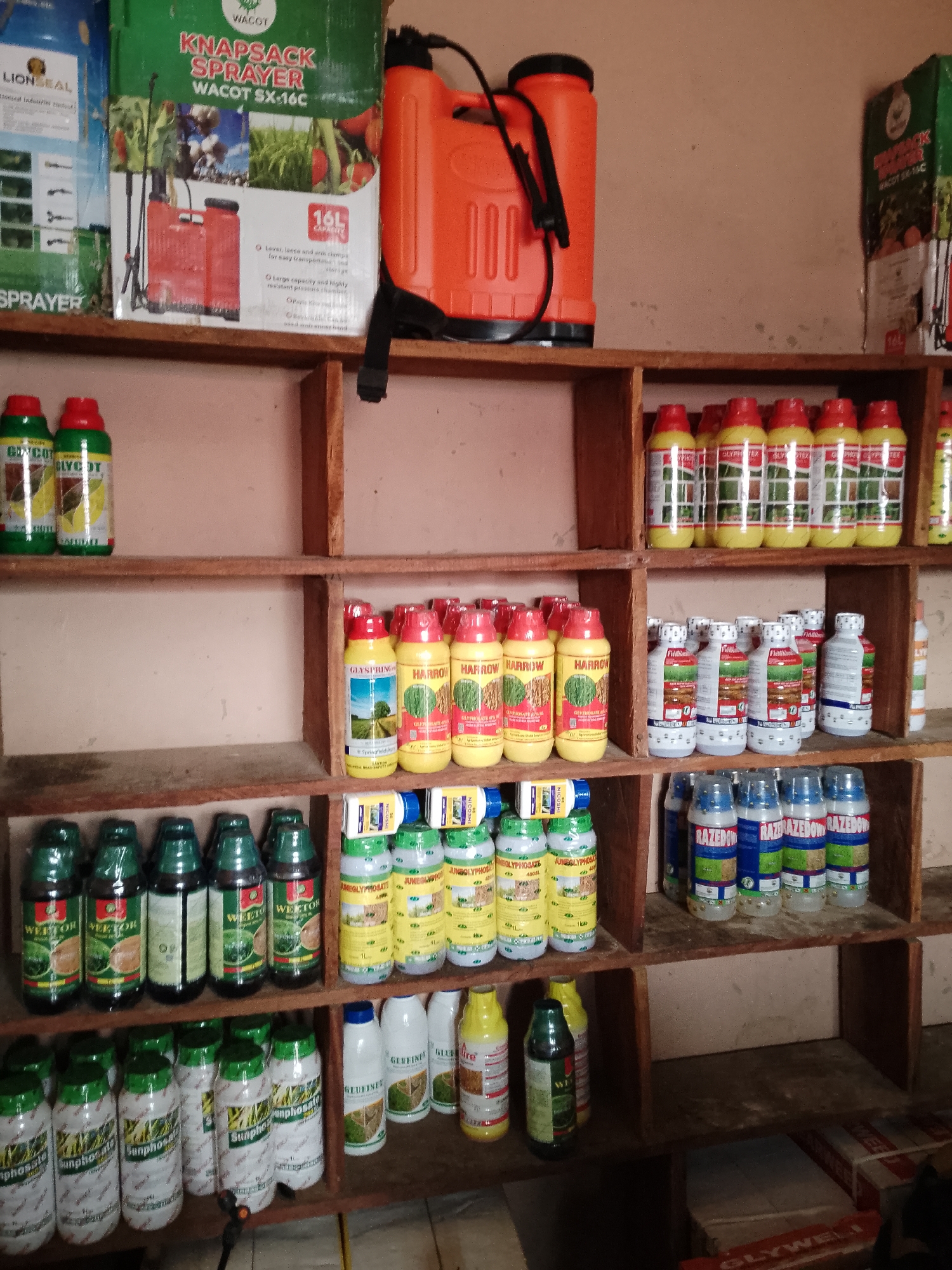 The image shows shelves stocked with agricultural chemicals, including sprayers, bottles of herbicides, and pesticides. On the top shelf, there is a red knapsack sprayer and boxes of agricultural products. The middle shelf is filled with rows of yellow and white bottles with red caps, and on the bottom shelf, there are green and white bottles. In the bottom right corner, there are boxes stacked. The scene appears to be a store or warehouse selling agricultural supplies.