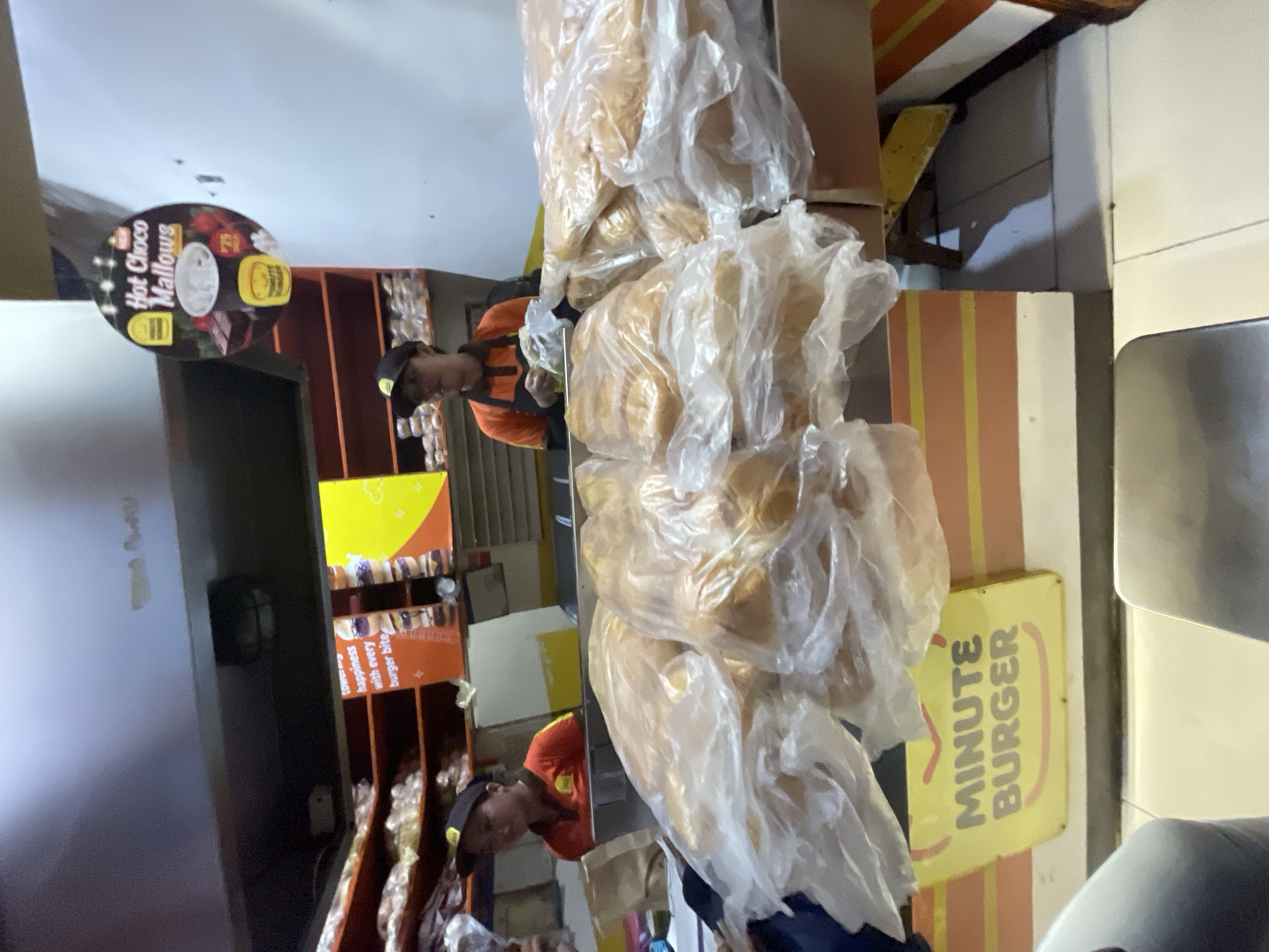 In Davao City, Philippines, a busy counter at a Minute Burger outlet is depicted. Two female employees, wearing orange shirts and caps, are visible behind the counter. One employee is in the process of serving a customer, holding a burger bun. Piles of burger buns, wrapped in plastic, are prominently displayed on the counter. Behind the employees, shelves are stocked with more packaged food items. A circular advertisement for "Hot Choco Mallows" hangs above the counter. A bright yellow sign with "MINUTE BURGER" written in gray letters is also visible. The scene captures the commercial activity and food service environment of the establishment.