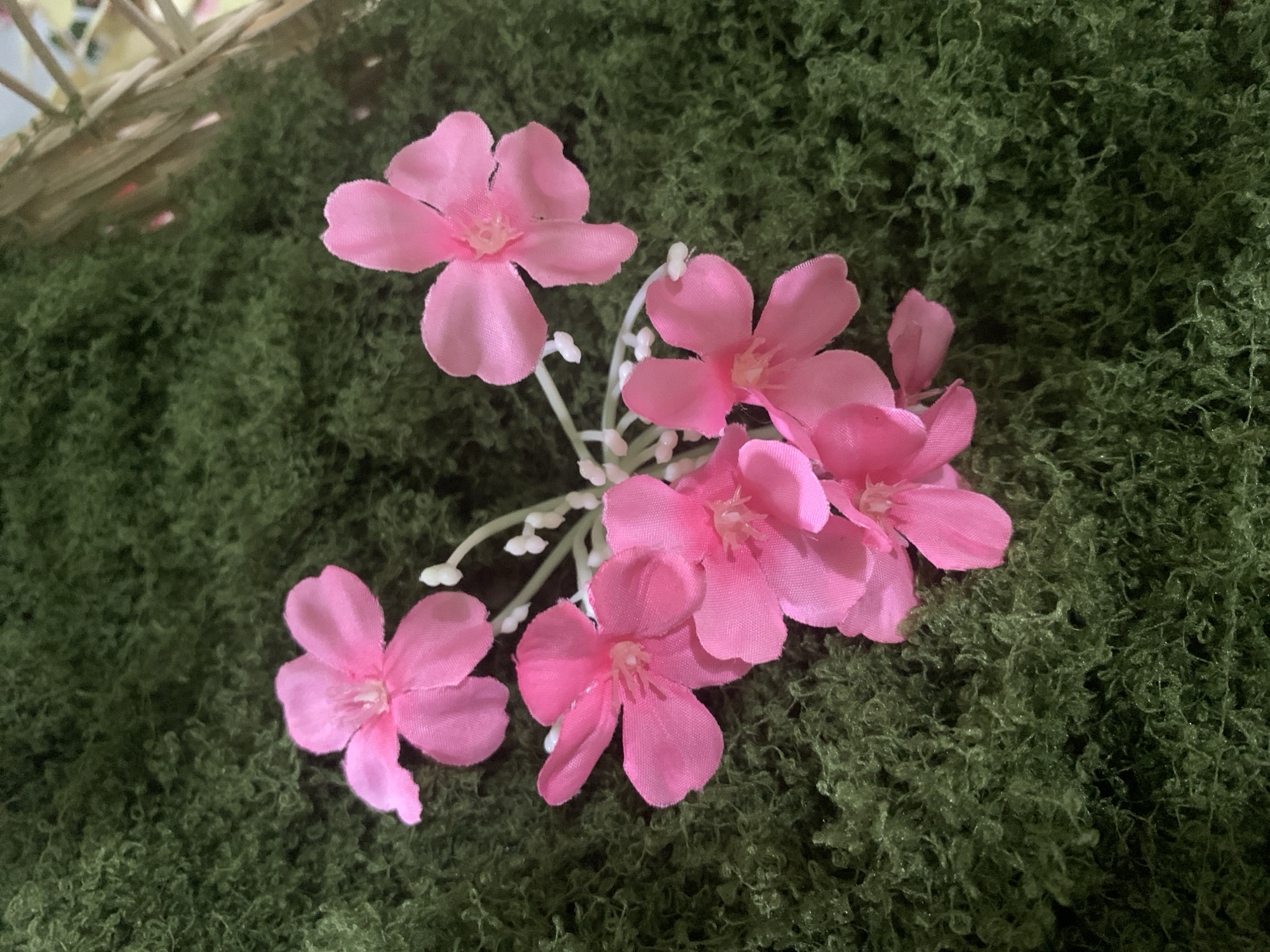The image shows a close-up, top-down view of a cluster of artificial pink flowers against a background of dark green, moss-like material. The flowers have five petals each and are arranged in a loose bunch, connected by thin, white stems and small white buds. The lighting is soft, creating subtle shadows that highlight the texture of the moss. The background moss is dense and uneven, with visible strands and clumps. In the upper right corner, a small section of a woven, light-colored basket is visible. The scene appears to be a still life arrangement, possibly for decoration. There are no people or animals present, and the setting does not provide any specific visual cues to pinpoint its location within Kuala Pembuang, Indonesia.