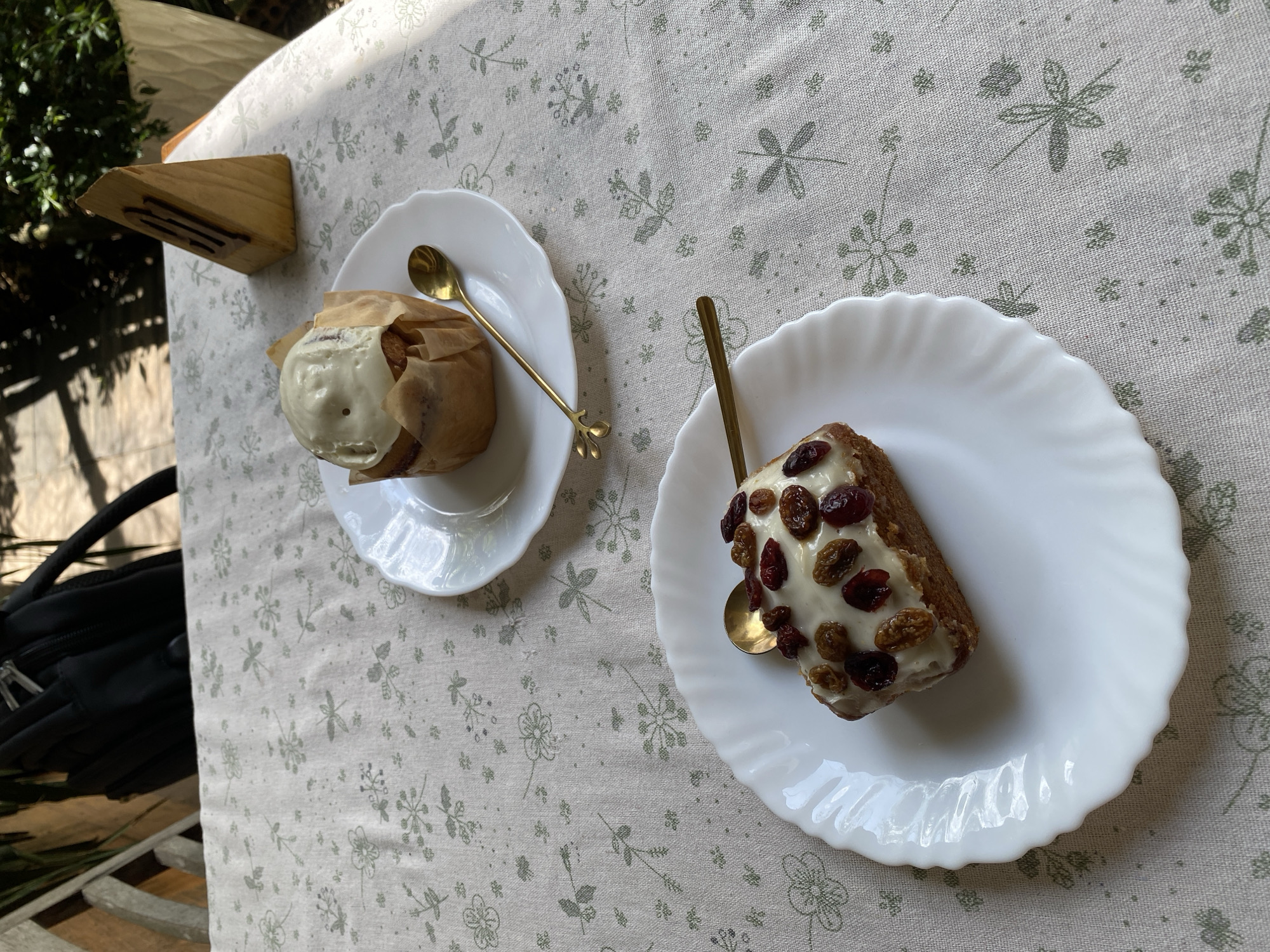 A close-up shot showcases two desserts on white plates with ruffled edges, placed on a light-colored tablecloth with a subtle green floral pattern. The dessert in the foreground is a rectangular cake topped with white icing and studded with dried cranberries and raisins. A small golden spoon rests on the cake. In the background, a round pastry, also topped with white icing, sits on its own plate. A similar golden spoon is positioned beside it. A triangular wooden object, possibly a menu holder, stands in the background to the right, partially obscured by greenery. The scene appears to be set outdoors, with dappled sunlight filtering through leaves, suggesting a café or restaurant setting in a city like Lâm Viên - Đà Lạt, Vietnam, known for its pleasant climate and natural beauty.
